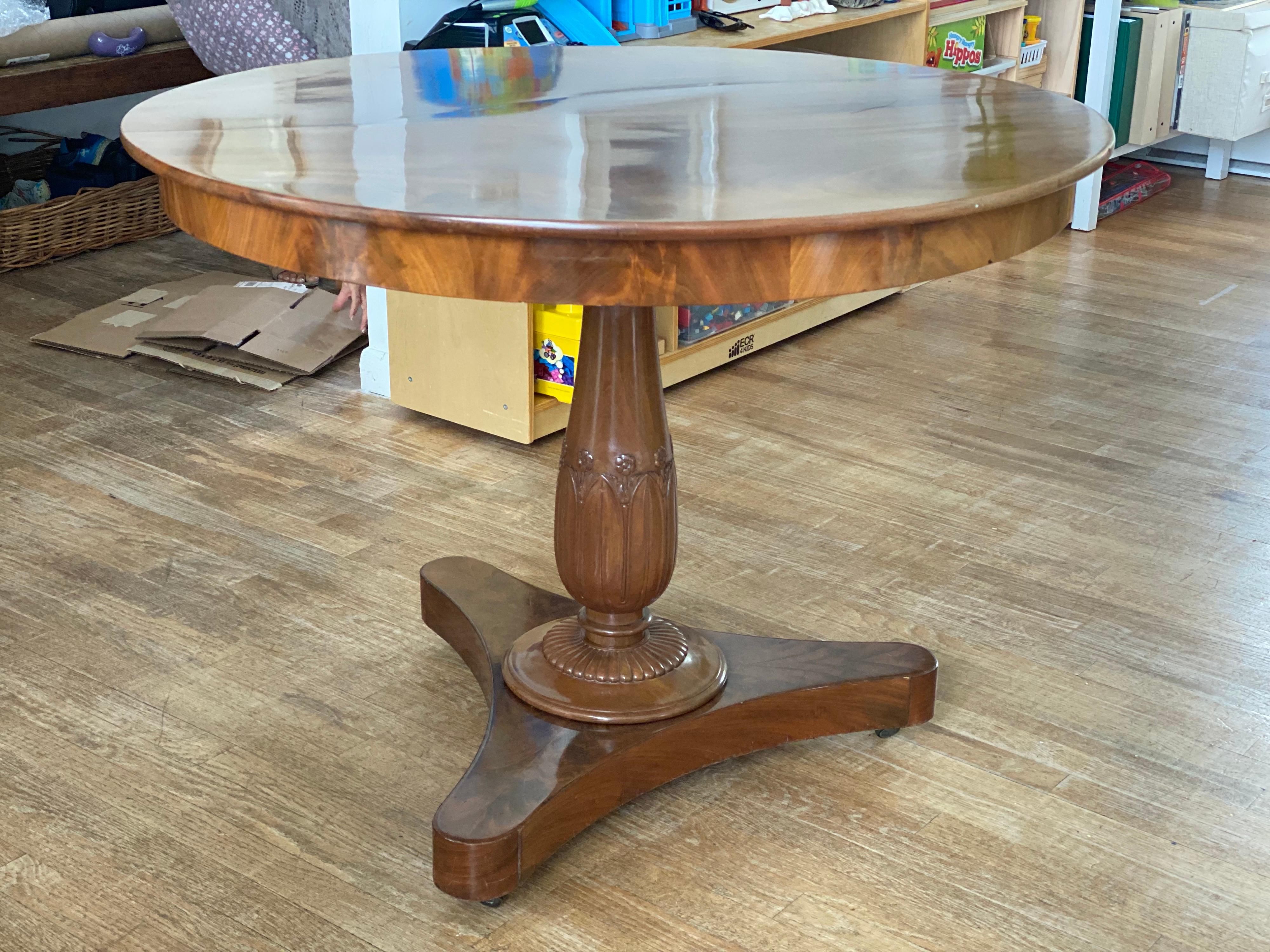 Round Mahogany Pedestal dining table, early 20th century
Beautiful flame mahogany top. Floral and leaf carving on pedestal ending on a tripod base on casters.
Some sun fading to top, light wear to finish, earlier restoration seen under top to stop