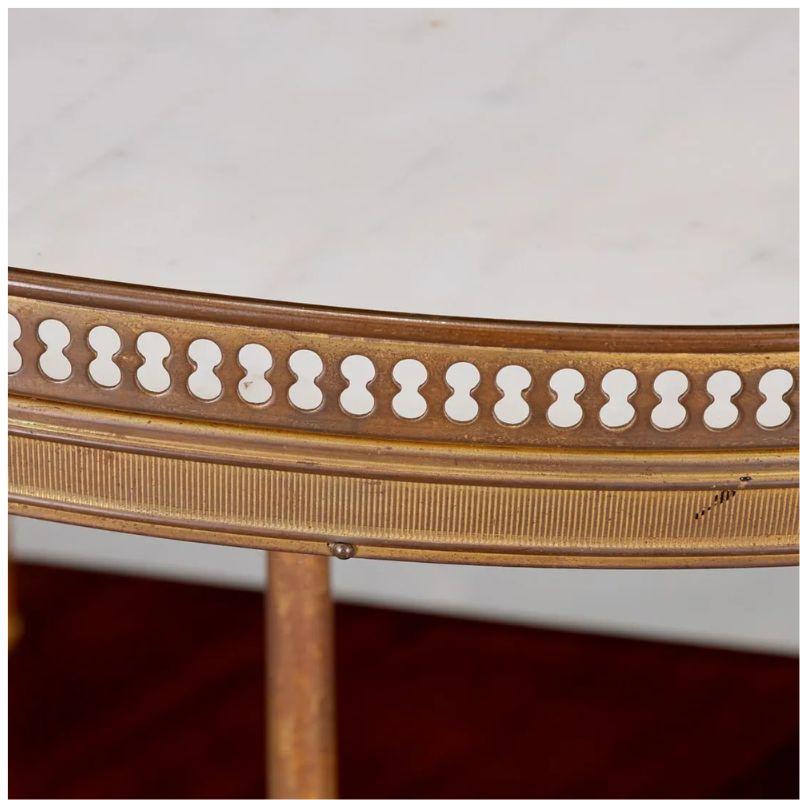 A vintage round marble top gueridon table with a brass rail, brass legs and mahogany shelves. At the heart of this gueridon table is a round white carrara marble top, exuding a luxurious allure that adds a touch of grandeur to any space. Encircling