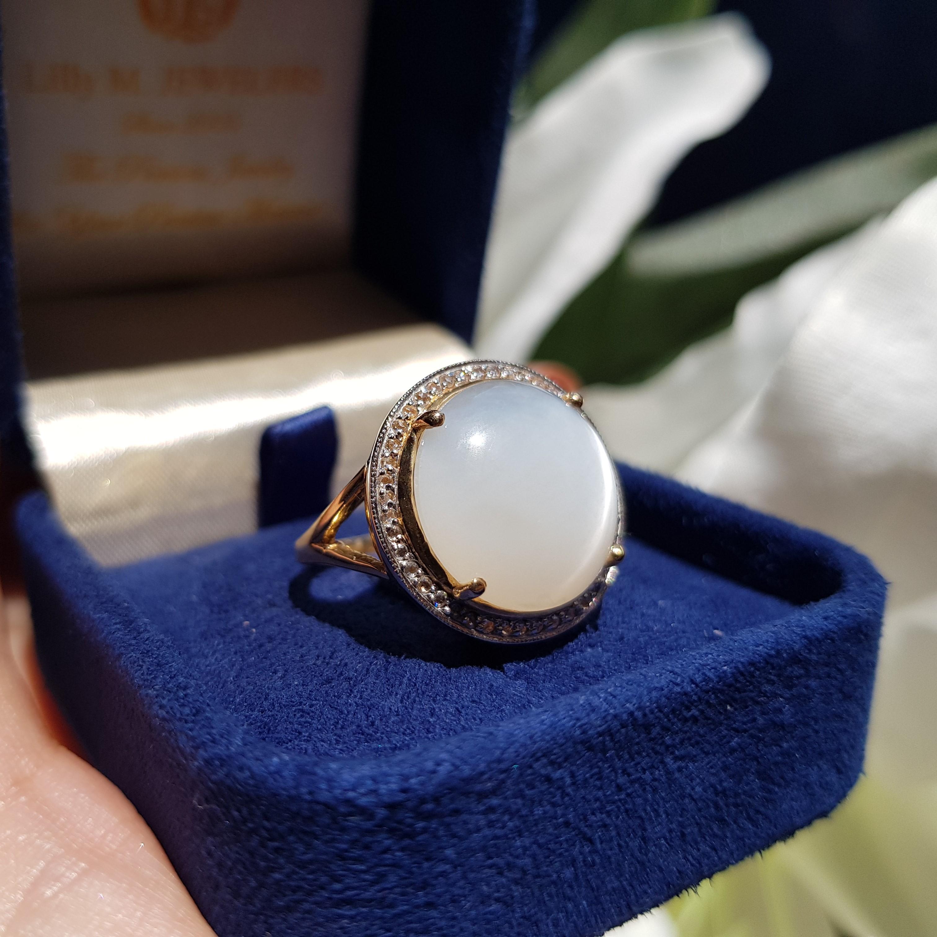 This stunning women’s fashion ring features a large round moonstone at the top, surrounded by a halo of round bright cut set diamonds, and perched atop a split yellow gold shank. The band is made of 9k yellow gold, and it’s softly rounded on top. It