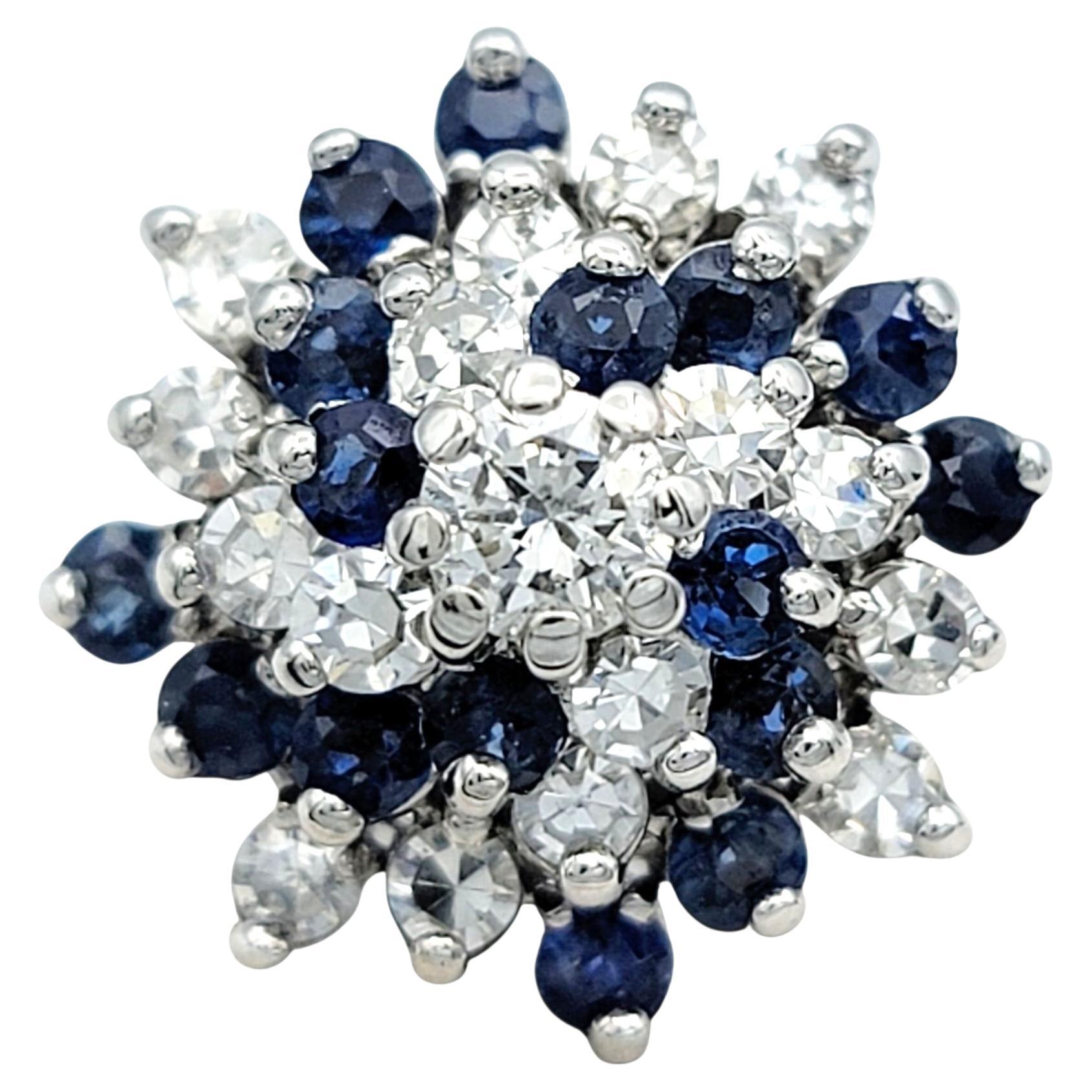 Dieser Clusterring aus 14 Karat Weißgold besticht durch eine Komposition aus natürlichen blauen Saphiren und Brillanten. In der Mitte verankert ein größerer runder Diamant das Stück, umgeben von abwechselnden Saphiren und Diamanten, die eine volle,