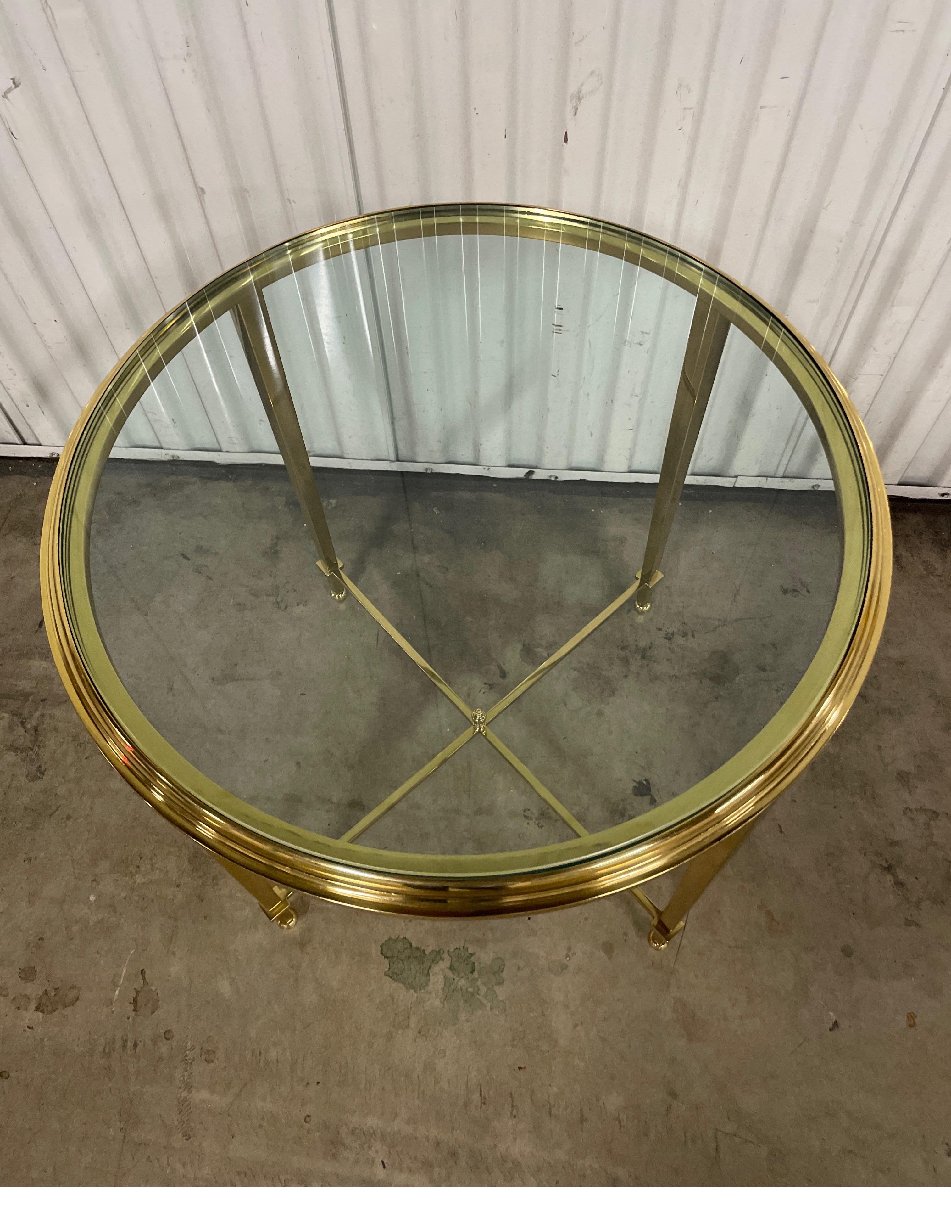 Vintage solid brass round side table with glass top by Jansen. The four legs are attached at bottom with an X stretcher and center finial. A very simple and elegant small drinks table.