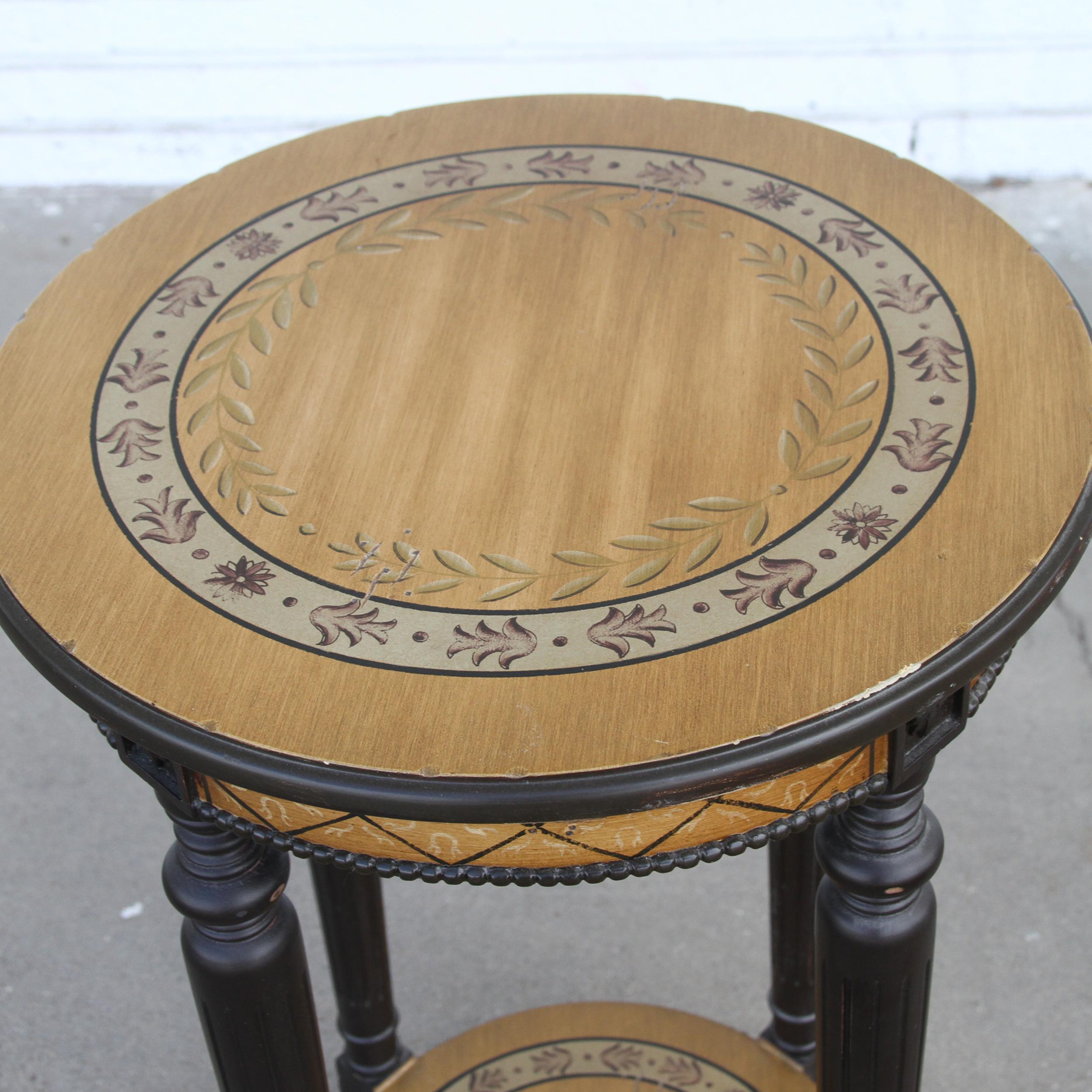Il s'agit d'une table d'appoint de style néoclassique avec un plateau rond. Il est peint de manière décorative et possède une étagère inférieure pour le rangement. Les pieds tournés sont peints en noir. La jupe et le haut sont décorés de peinture et