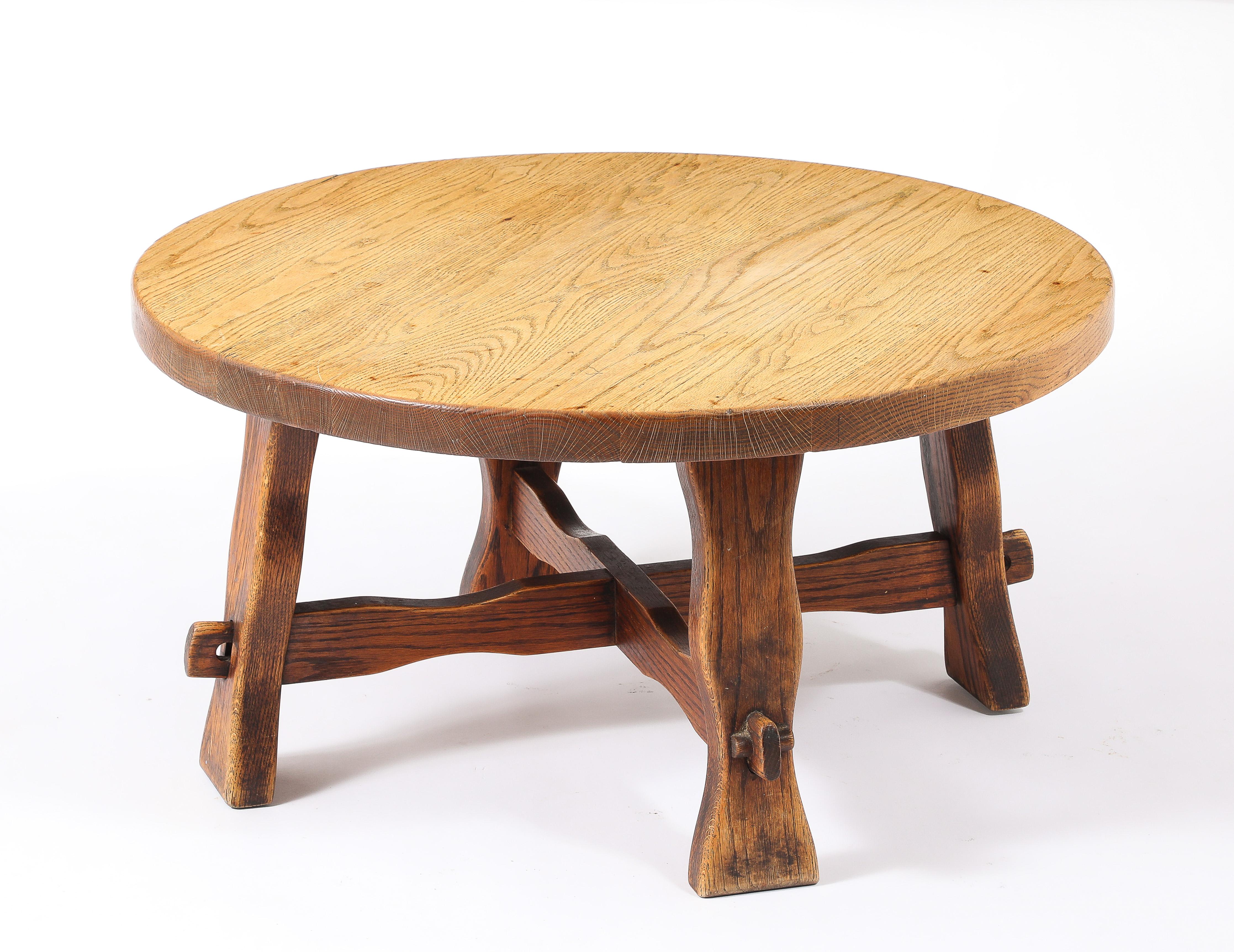 A round oak coffee table with a thick top on carved legs, joined by a carved stretcher and held together with wooden keys.