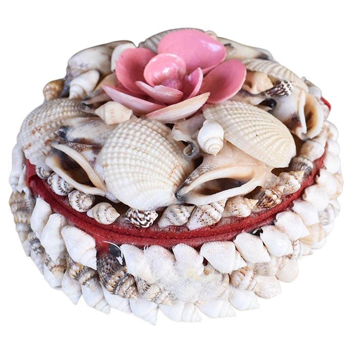 A pretty coastal-inspired round decorative box encrusted with a sea shell motif. This box is round and features an attached lid. The lid is decorated with an array of seashells and topped with pink painted shells arranged in a flower motif. The lid