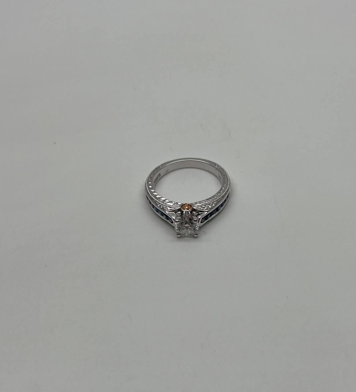 This exquisite piece features a brilliant central diamond set in a sleek, modern design. The band is crafted from polished white gold, showcasing intricate detailing along the shoulders. A small, round accent diamonds and sapphires adds a unique