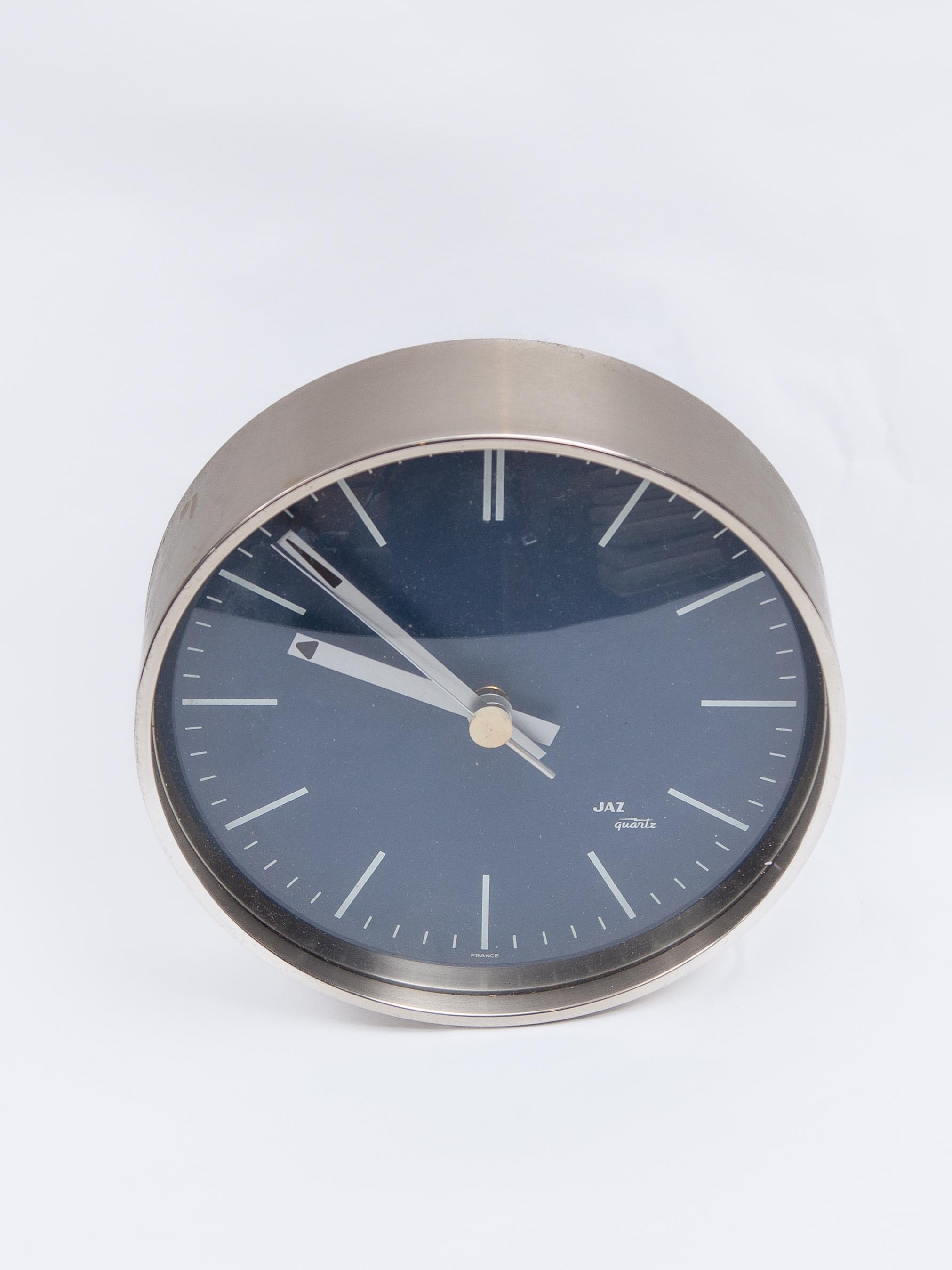 The Jaz Quartz is a round wall clock. The clock has a silver case made of brushed aluminium, a metal blue index. The aluminum dials give this clock a lovely chic and stylish design that fits in many interiors. This clock is silent and works on 1 AA