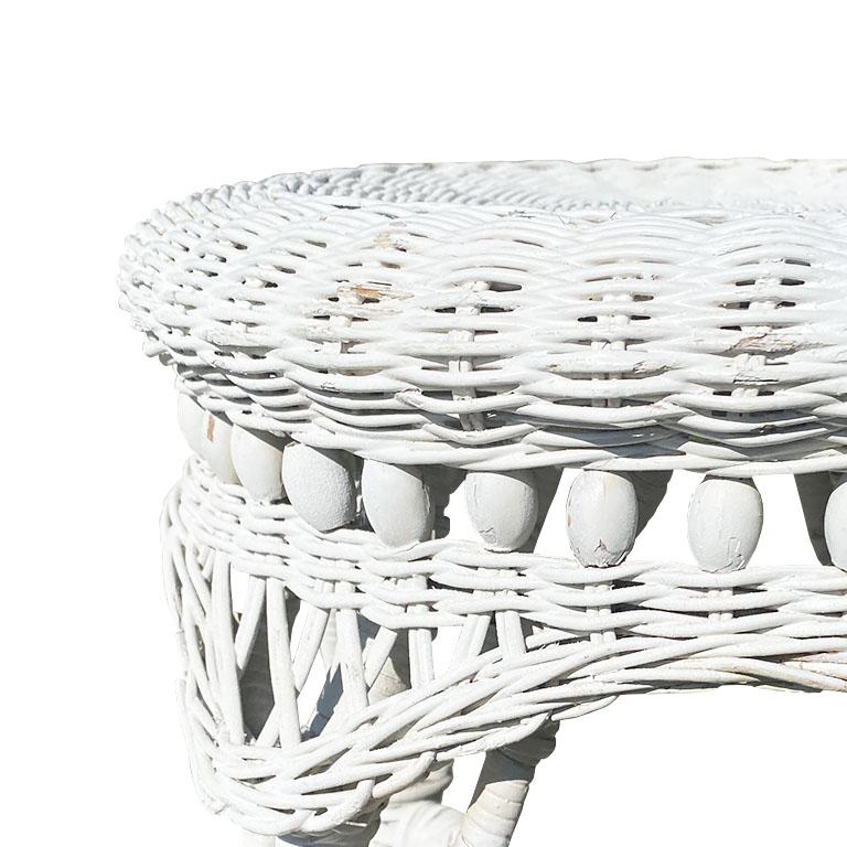 A white vintage wicker plant stand or stool in the style of and Heywood-Wakefield. This piece features a woven round wicker top that would be perfect for use as a plant stand. Its four splay legs are wrapped in rattan and feature an X stretcher at