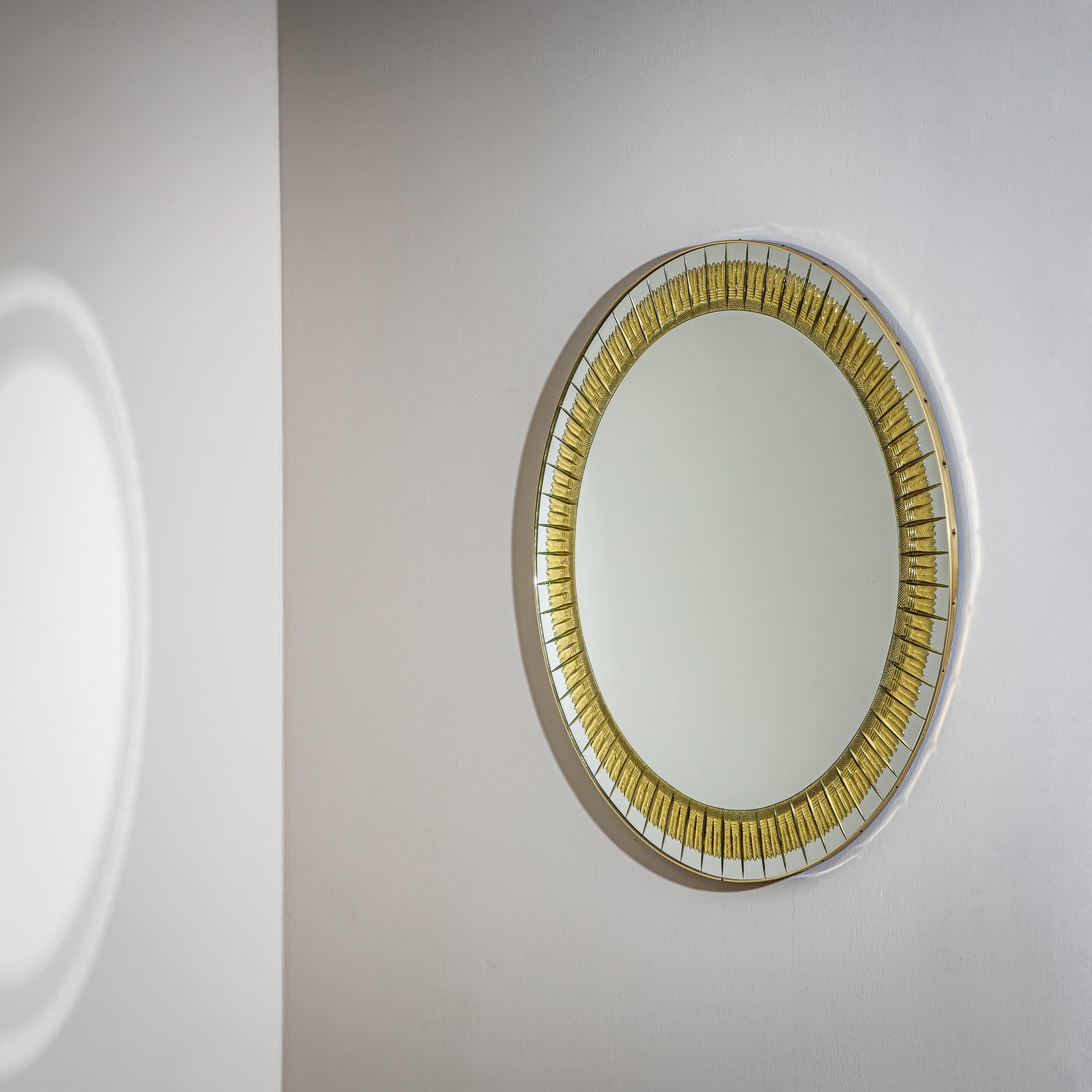 Miroir mural rond avec un cadre plat en laiton, entouré de segments de miroir à facettes individuels disposés en cercle. Un motif solaire doré en forme de plumes stylisées rayonne vers l'extérieur.