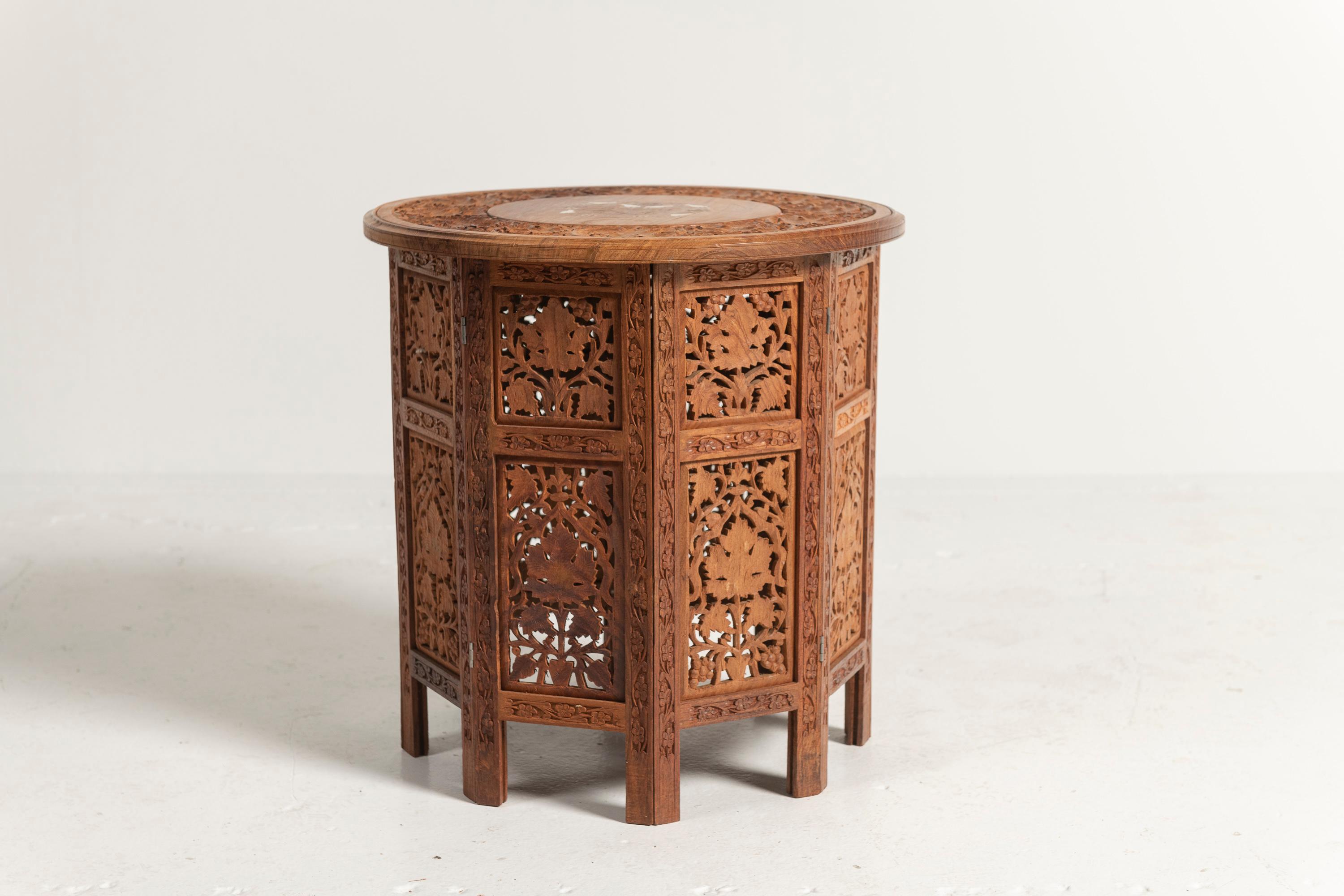 Este taburete o mesa auxiliar redonda de teca marroquí irradia una artesanía intrincada. El tablero de la mesa presenta un bello motivo de pájaro tallado con delicadas incrustaciones de hueso, que añaden un toque de elegancia. La base octogonal está
