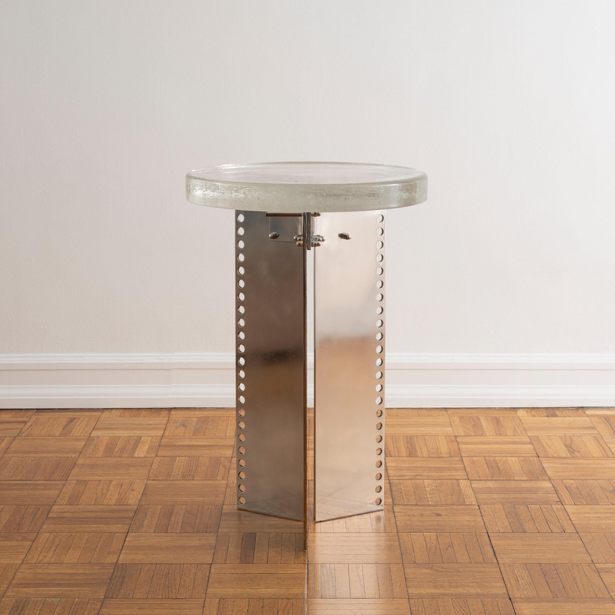 Contemporary accent table comprised of a round glass top fixed to an architectural base of perforated stainless steel. 

The glass is produced by crucible casting, which creates an organic rippled texture on the underside and features natural chill