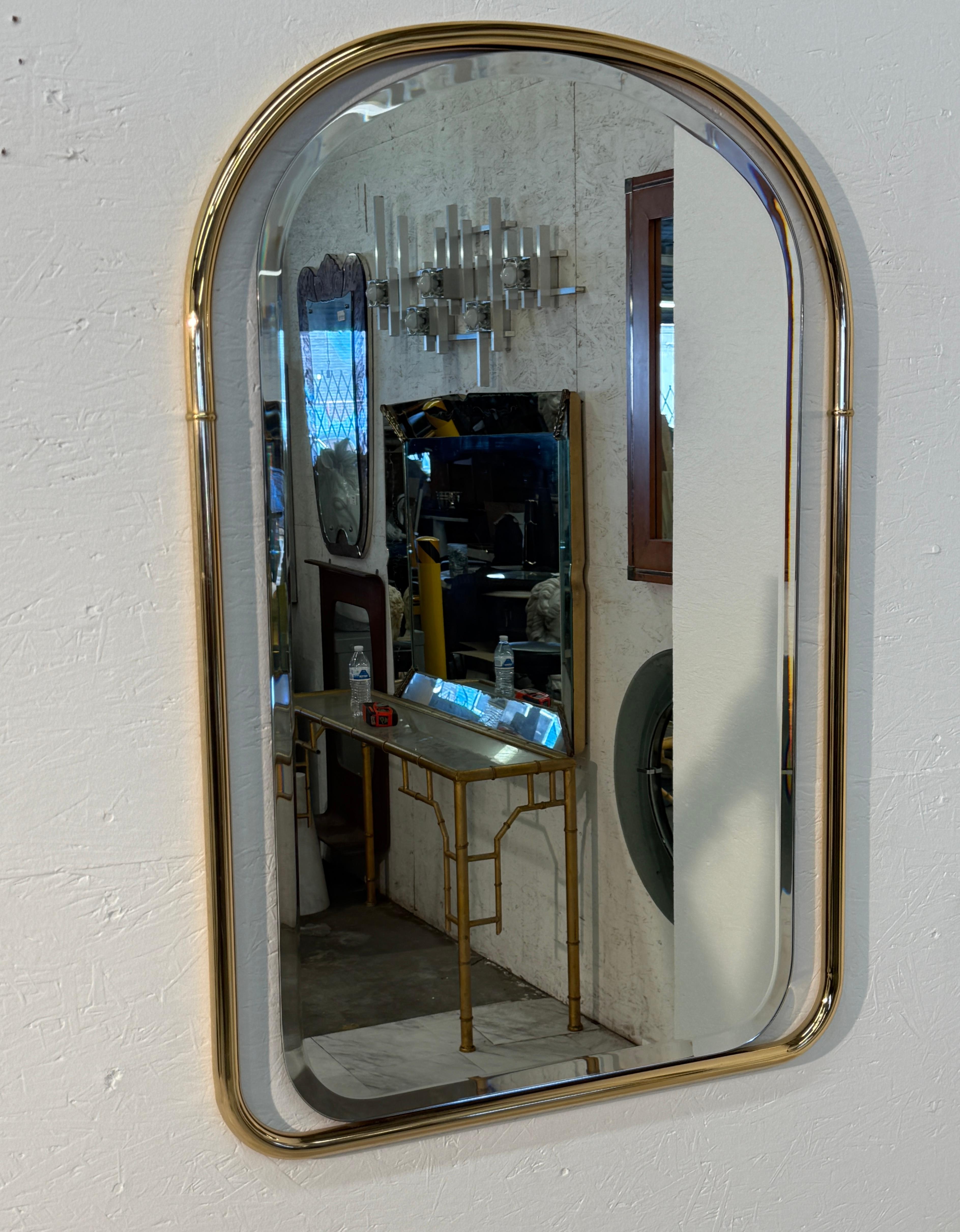 Espejo de pared de estilo vintage con marco de latón pulido, parte superior redondeada y bordes curvados. El armazón tiene un diseño tubular liso con un sutil acento anular en la línea media a cada lado, que añade un refinado detalle decorativo. El