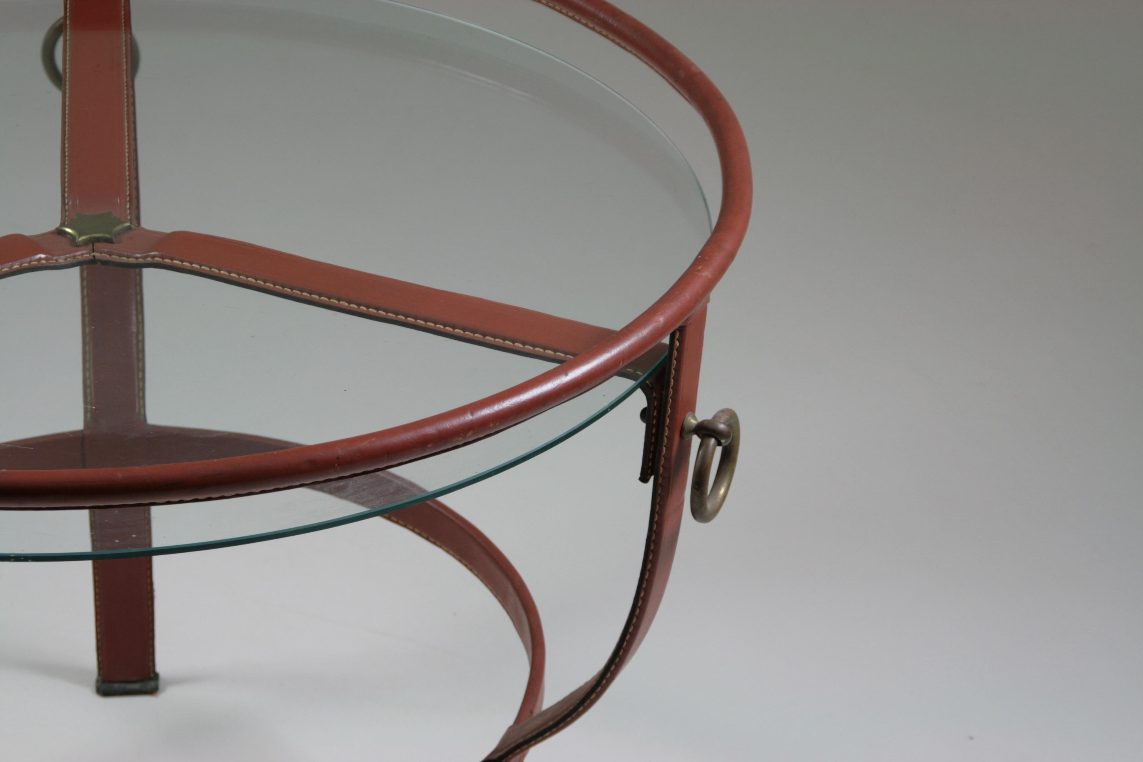 Seltener und eleganter runder Dreibein-Couchtisch von Jacques Adnet. Rohrförmige Metallstruktur, vollständig mit braunem Sattelleder bezogen und mit einer Glasplatte versehen. Sehr guter Zustand. Seltene Kratzer vom Gebrauch auf dem