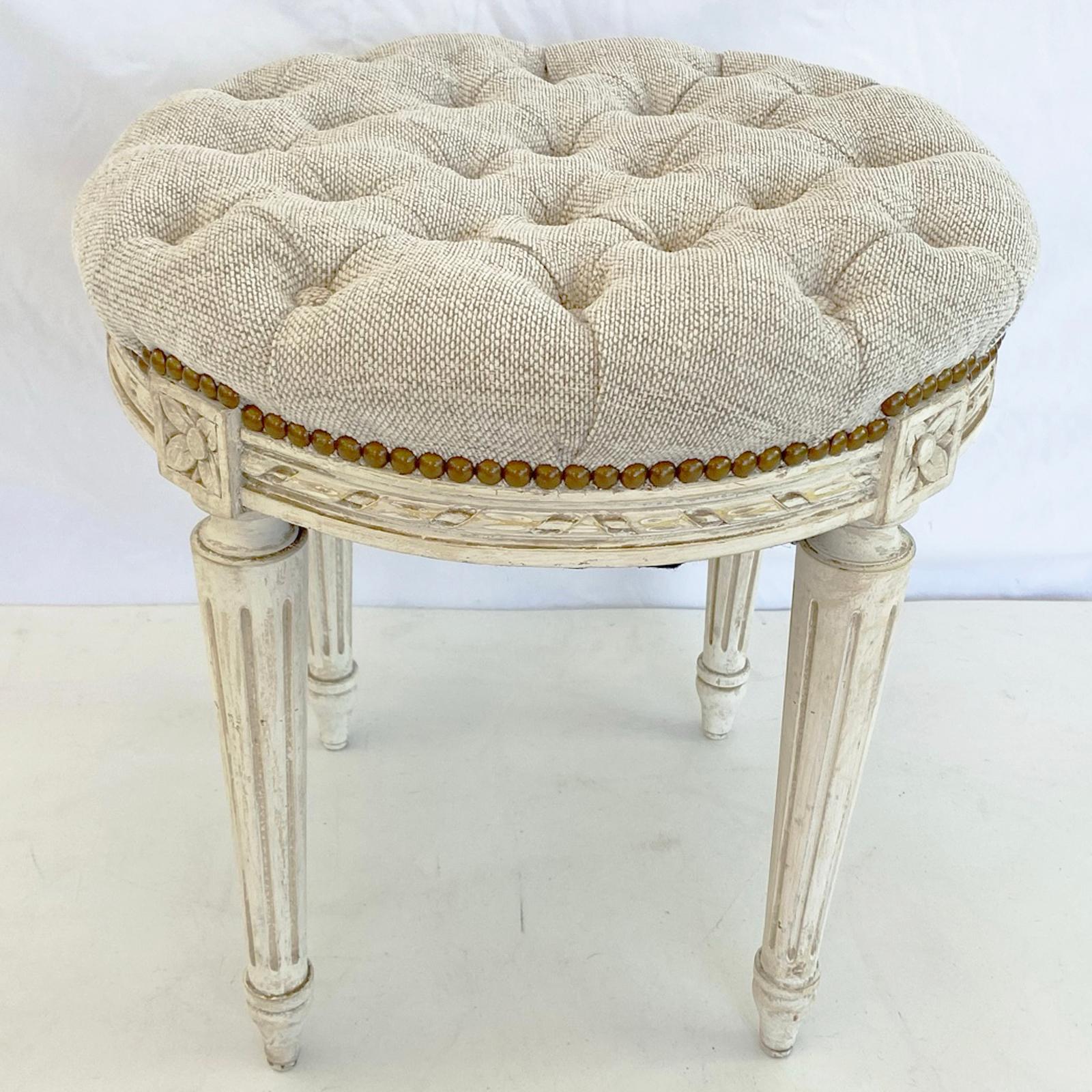 Round stool, having a tufted crown seat upholstered in twill fabric with nailheads, on a painted base, its apron carved with ribbon gadrooning, rosette-blocks over four round, tapering, fluted legs, ending in touipe feet. 

Stock ID: D3050.
