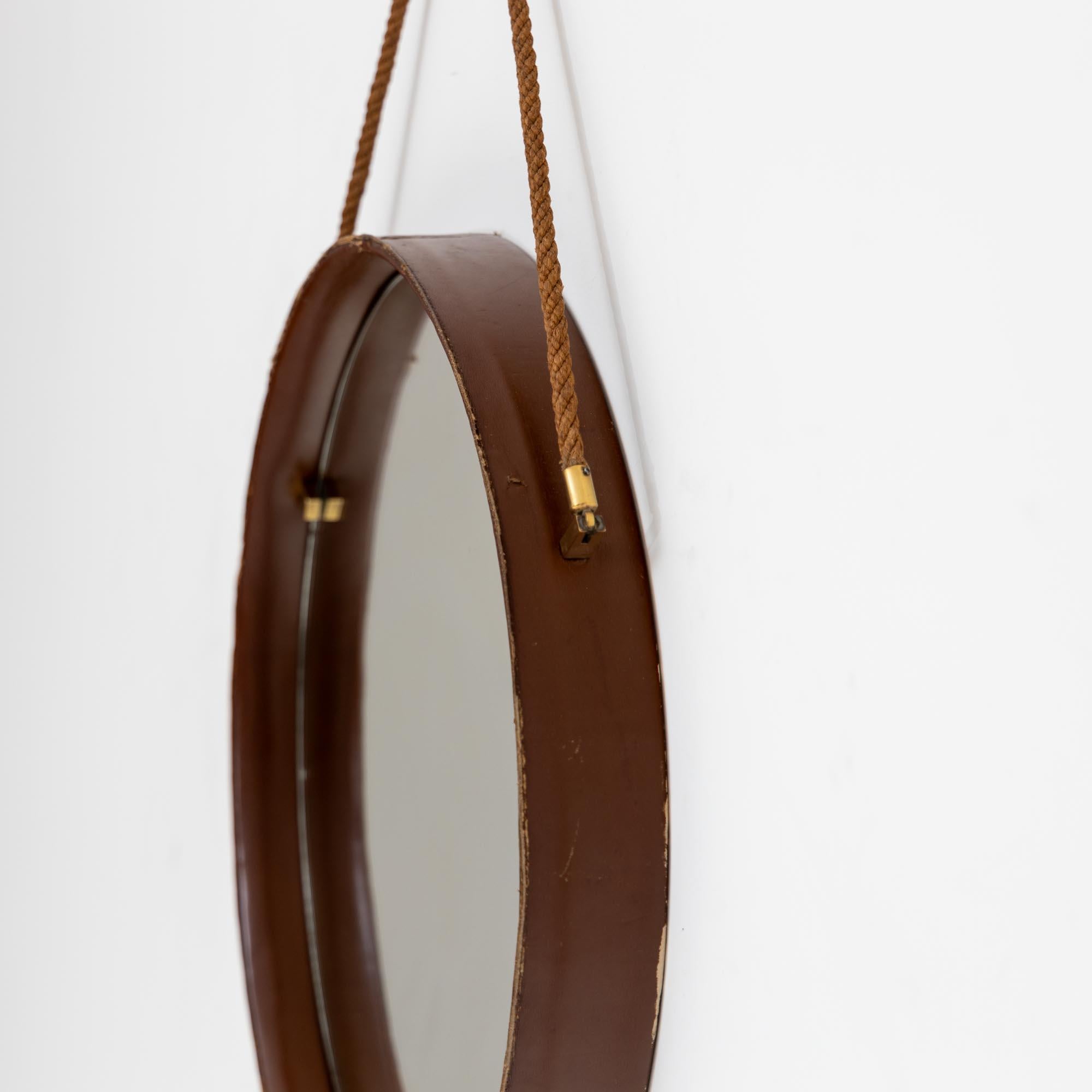 Round wall mirror with high rim with dark brown leather covering and short brass mountings. The mirror hangs on a short rope which is fixed at two points on the outer frame.