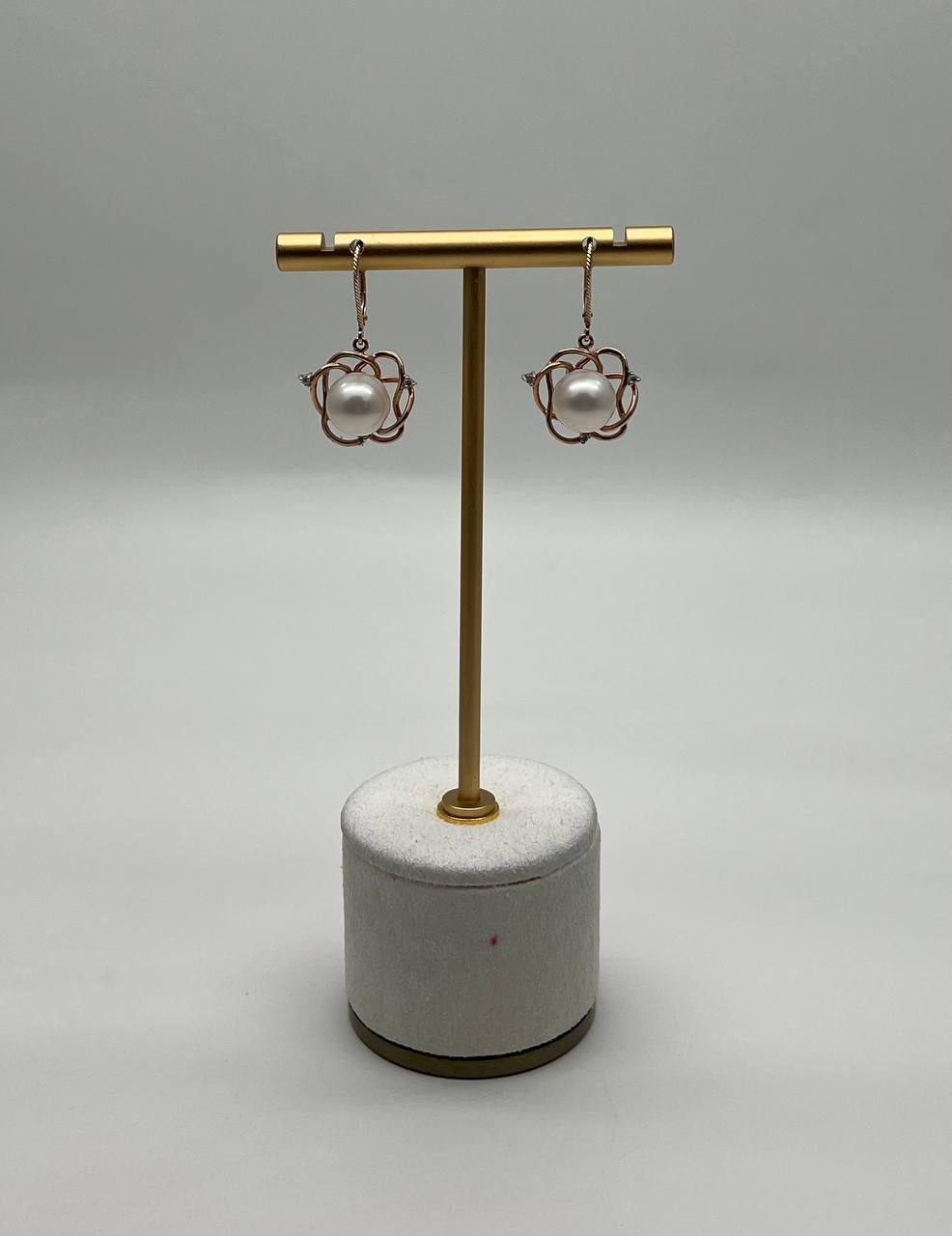 A pair of earrings, featuring a spherical white pearl encased in an intricate solid gold wire design. The earrings are suspended from a twisted gold hook, offering a modern aesthetic. 14K Gold. Natural Pearls.