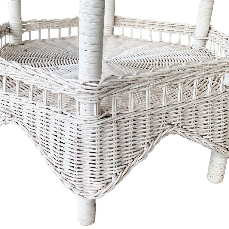 A gorgeous white woven wicker side table with a bottom shelf. The top of this occasional table is round, with edges that curl downward. Its octagonal base has wrapped rattan or bamboo legs that attach to an octagonal bottom shelf for extra storage.
