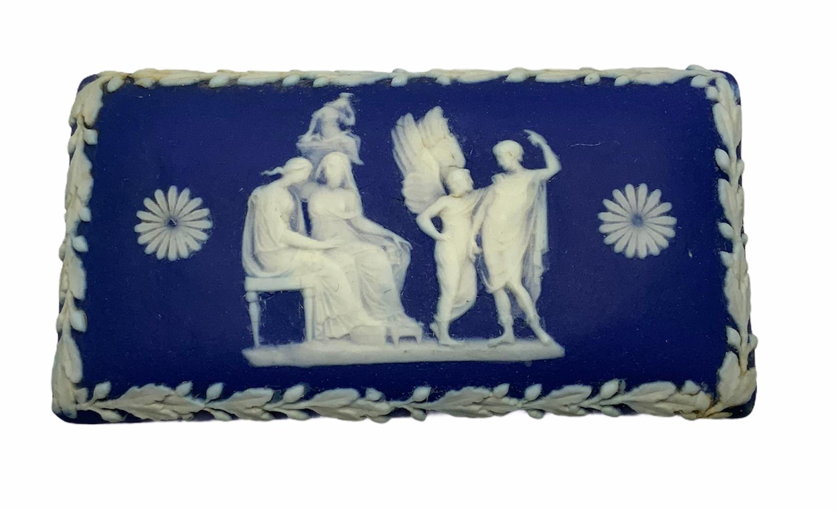 This is a rectangular jasperware trinket box depicting a scene with a white relief technique of two Greek or Roman ladies sitting in a settee while another lady is over them in heaven. In the same scene, an angel appears in front of them talking