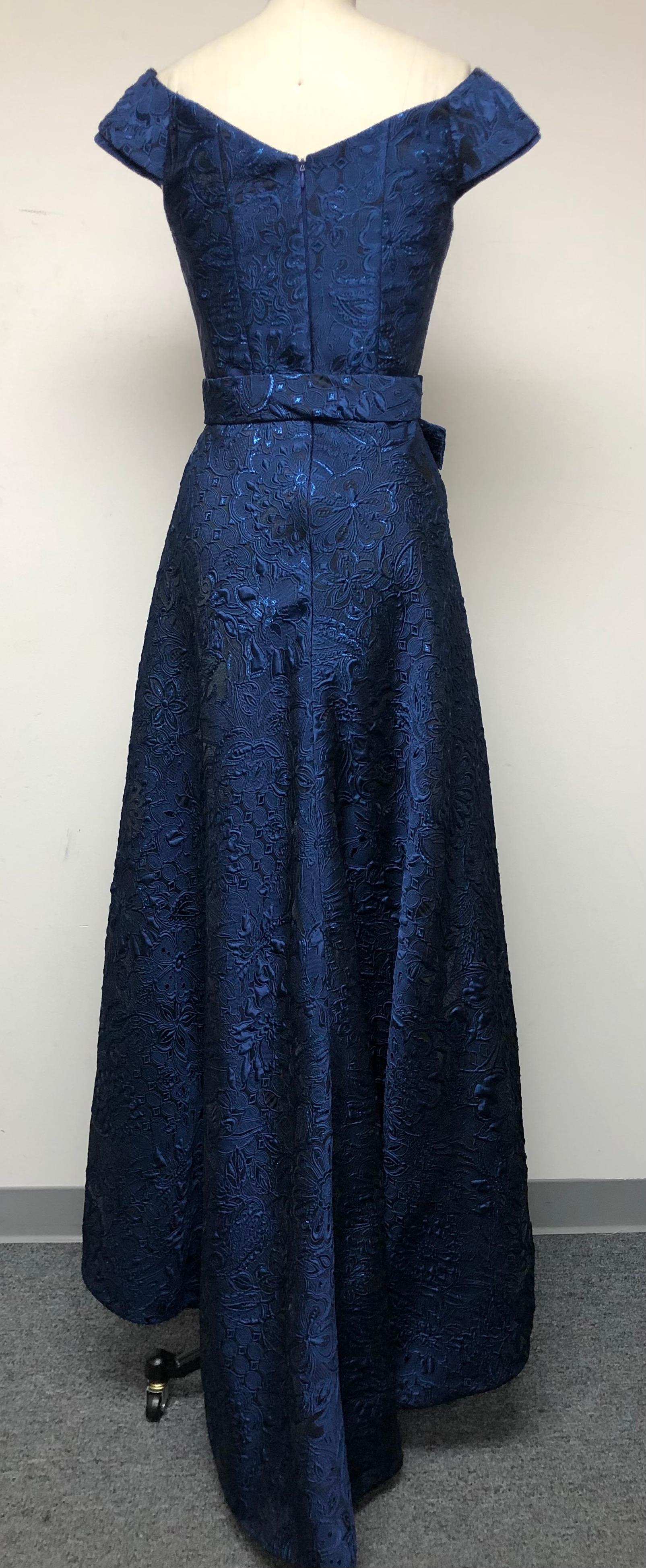 Navy blue portrait V neck fit flare high low gown. Cinched at the waist with self bow belt and chic train. In blue French matelassé with shimmering blue lurex for a bit of sparkle. Holiday dressing at its finest. For a sweeping entrance to parties.