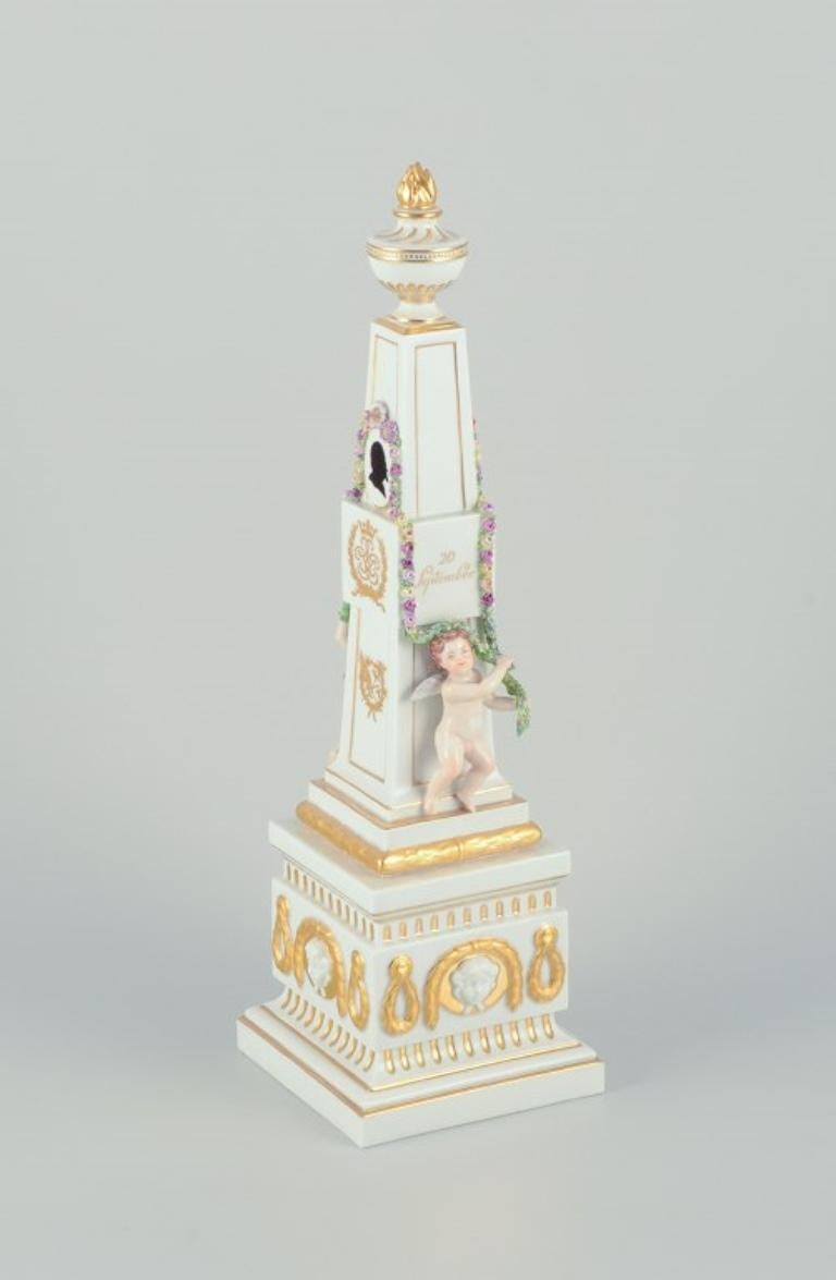 Royal Copenhagen Flora Danica.
Porcelain obelisk for table decoration.
Putti surrounded by garlands, budding flowers. 
24-karat gold leaf decoration.
Royal monograms 1884-1909 silver wedding anniversary.
Juliane Marie mark.
Approximately from the