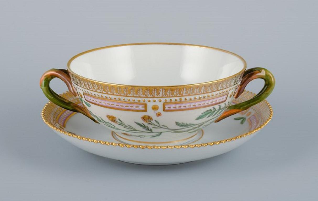 Royal Copenhagen Flora Danica Bouillontasse mit Untertasse aus handbemaltem Porzellan mit zweigförmigen Henkeln, Blumen und Golddekor. 
HERKUNFT: SCHLOSS VALDEMARS
Modellnummer 20/3612. 
Maße der Tasse: 13 x 5,5 cm.
Durchmesser der Untertasse: