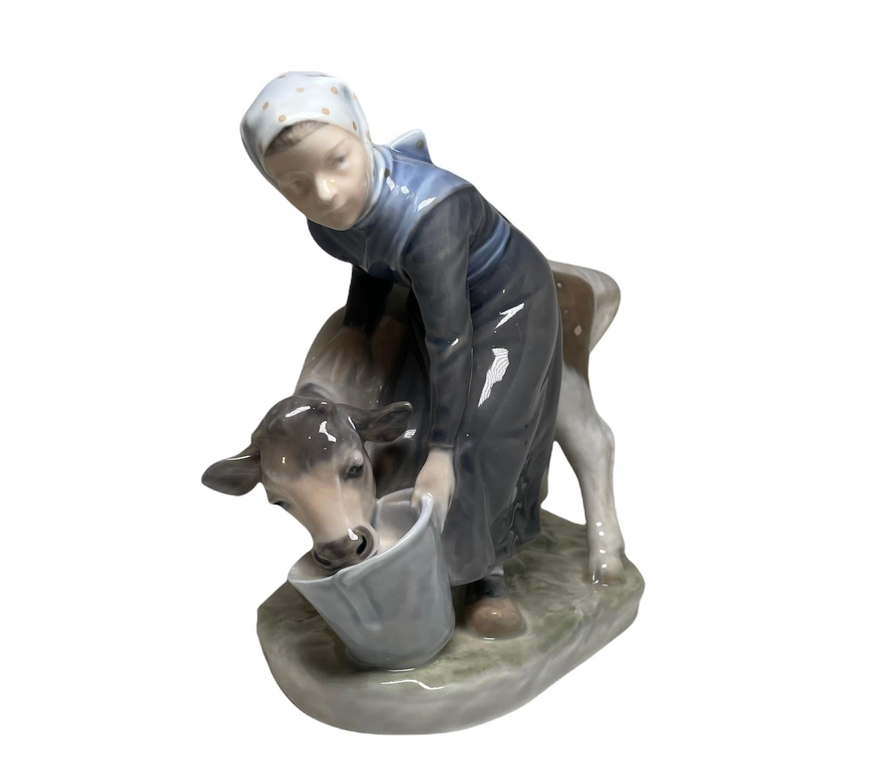 This is a Royal Copenhagen glazed hand painted porcelain figurine of a Danish young girl peasant feeding a calf from a big bucket. Below the base, it is found the Royal Copenhagen hallmark.