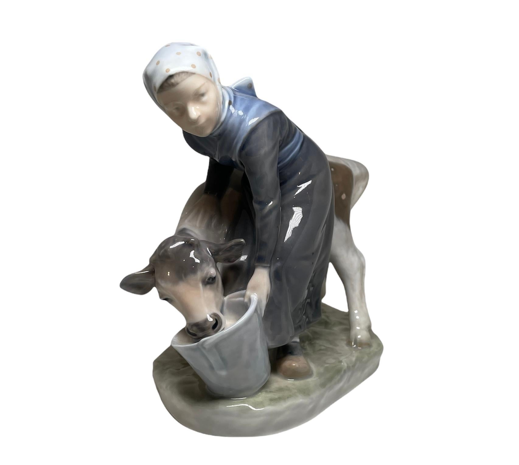 Questa è una statuetta in porcellana smaltata e dipinta a mano di Royal Copenhagen raffigurante una giovane contadina danese che nutre un vitello da un grande secchio. Sotto la base, si trova il marchio Royal Copenhagen.