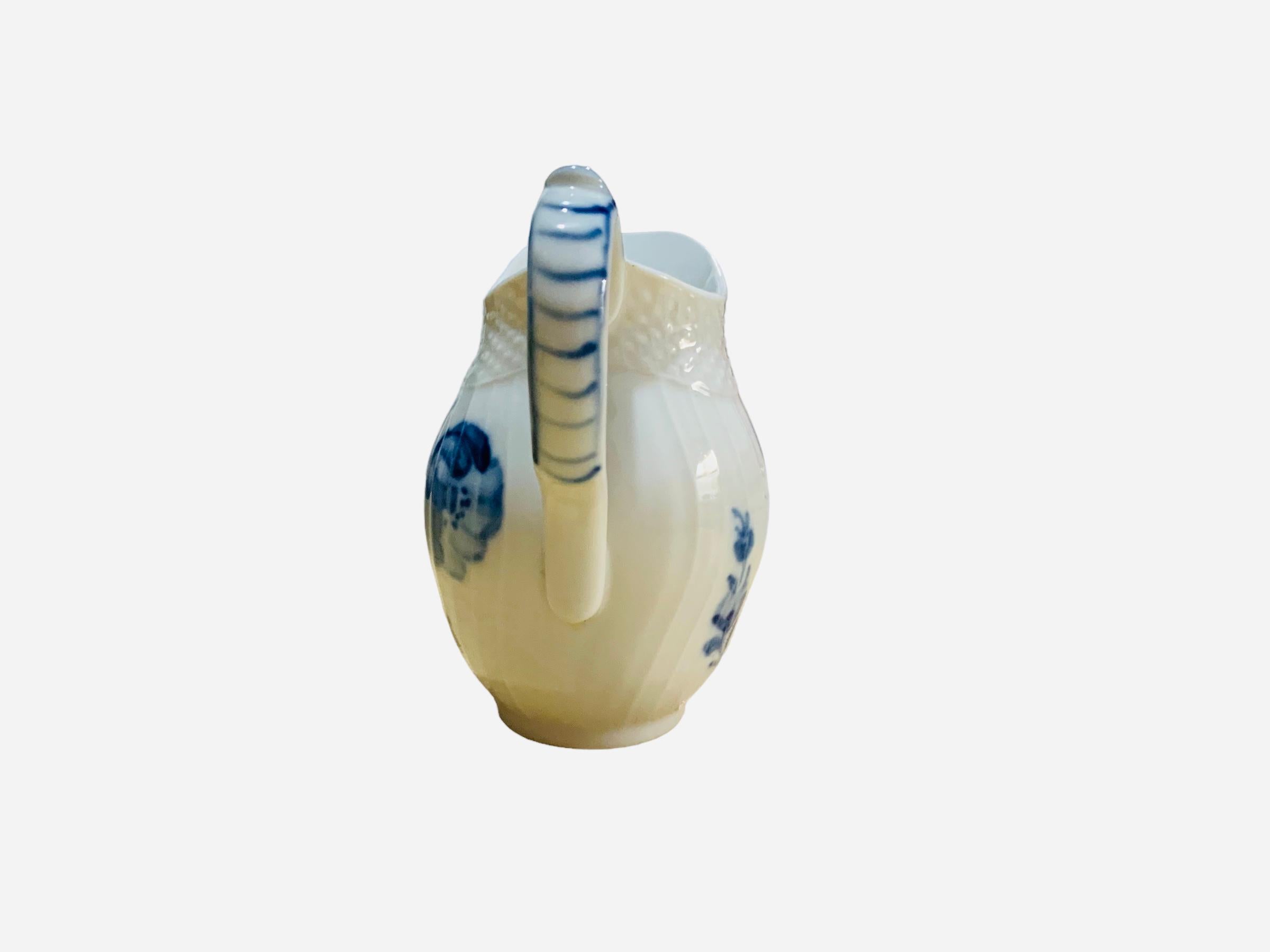 This is a Royal Copenhagen porcelain creamer. It has a white background and it is hand painted with a blue bouquet of flower in the center and single branches around the body and in the handle. The outside border of the small jar upper top is