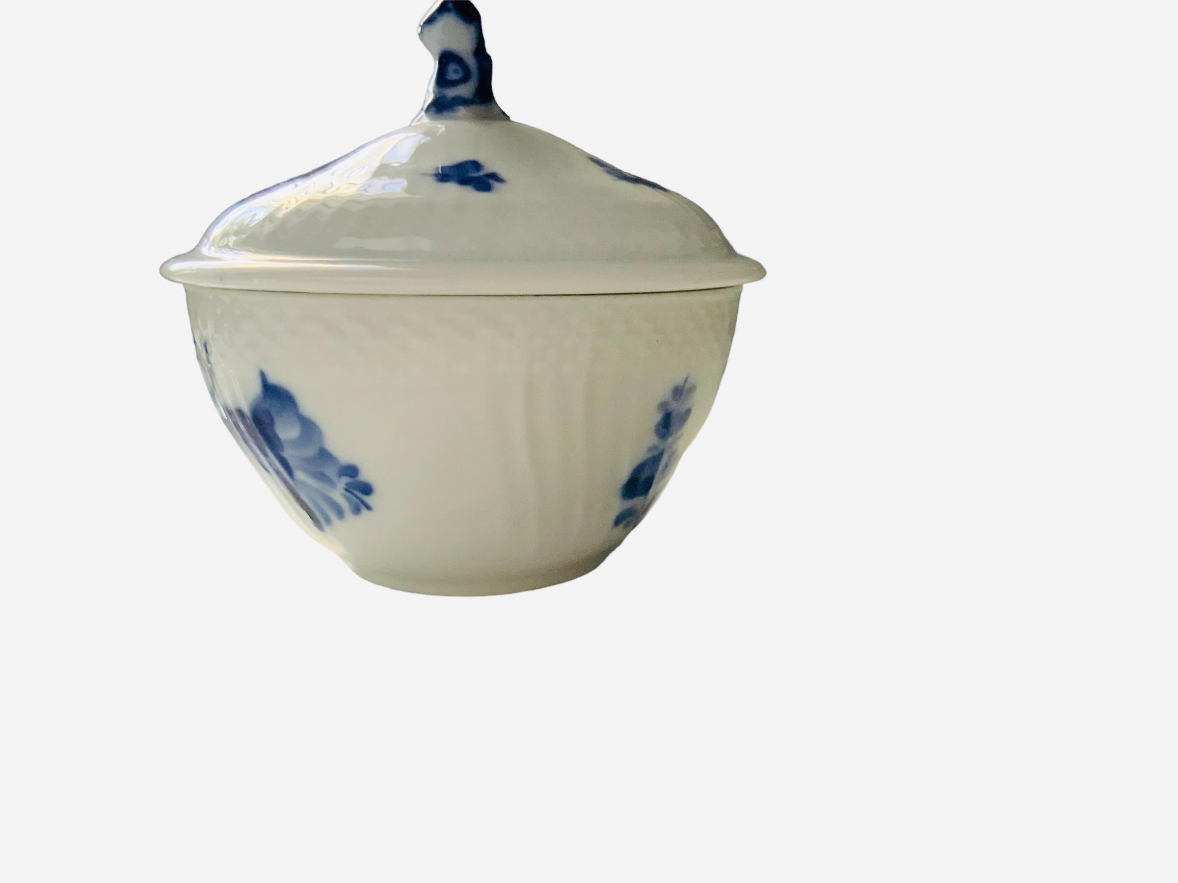This is a Royal Copenhagen porcelain sugar bowl with lid. It has a white background and it is hand painted with a blue bouquet of flowers in the center and single branches around the body and lid. The outside border of the upper top bowl and lid are