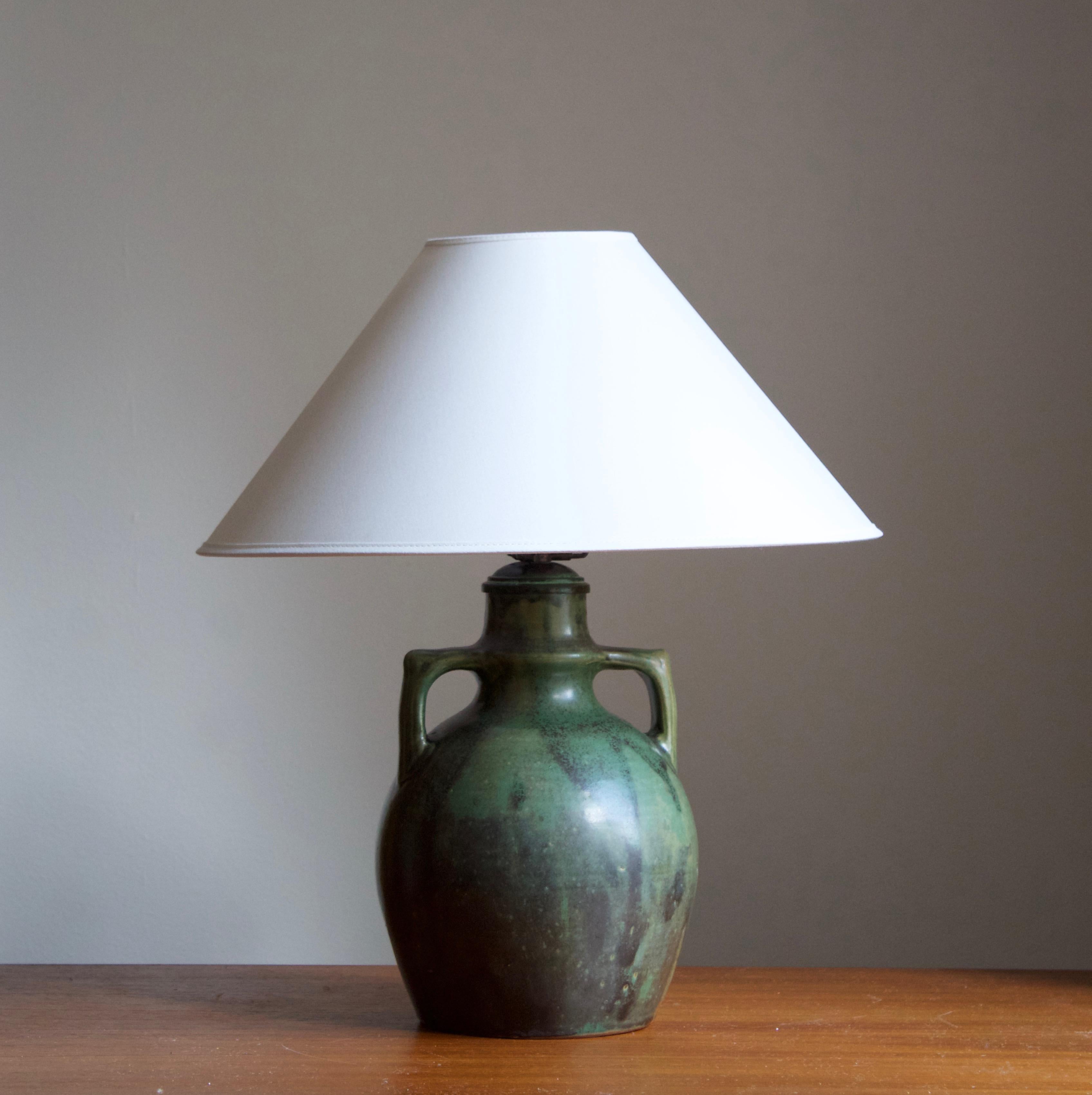 A table lamp designed and produced Royal Copenhagen, Denmark, dated 1927. Marked. 

Sold without lampshade. Stated dimensions exclude lampshade, height includes socket. Excludes remains of harp.

Glaze features a green color.