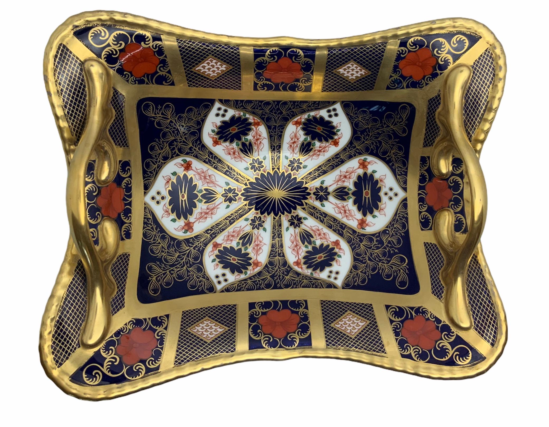 This is a Royal Crown Derby hand painted porcelain basket shaped as a concave parallelogram supported by a round base. It is hand painted in bright red, black, white and 22 carat gold. In the rectangular center predominates a large flower with a
