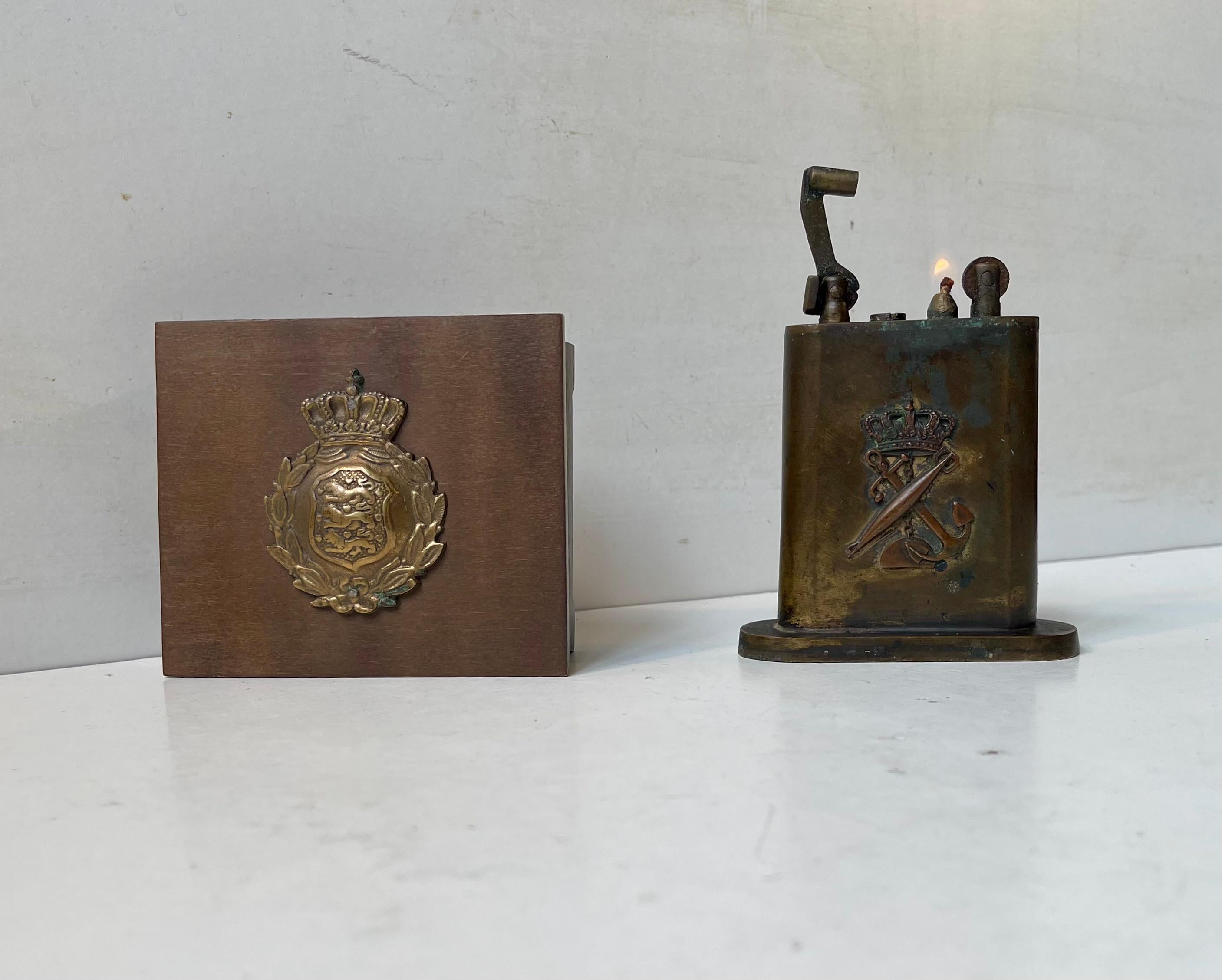 A Royal Danish Military set consisting of standing petrol lighter with the ciffers/code of arms of The Royal Danish Navy (Søværnet) and a cigarette box with the M23 Steel Helmet logo of the Danish brigade. Both items has a military a unproven
