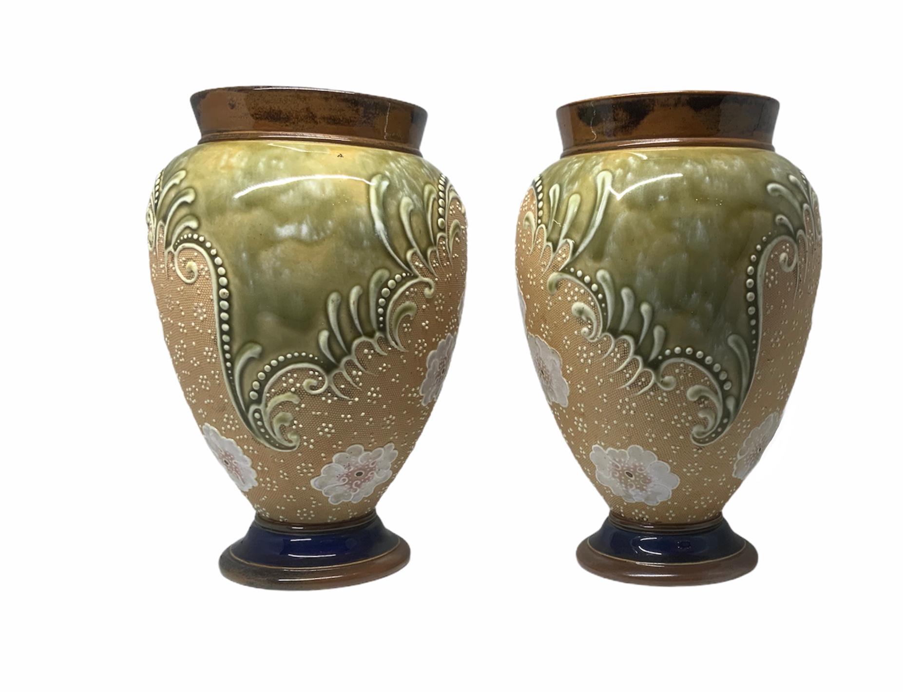 These are a pair of Doulton and Slater hand painted stoneware Amphora shaped vases. Their background is a combination of brown, green and, beige colors. They are adorned with a group of white flowers, dots all around, and framed by scrolls. Below