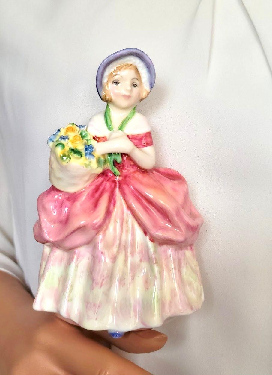 A lovely miniature vintage porcelain figurine Royal Doulton “Cissie,” HN 1809, decorated in a light pink colorway, was designed by leading Doulton artist Leslie Harradine in the “Harradine – Child Classics” Series. Even the basket of flowers on her