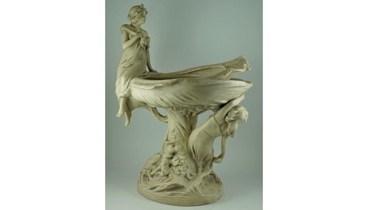 A bowl in the form of a shell, at which two water nymphs play - one hid under it, the other sat on its edge. The shell emerges from the waves, supported by the winding aquatic plants.
Royal Dux Bohemia, signature used until 1919.

Royal Dux is a