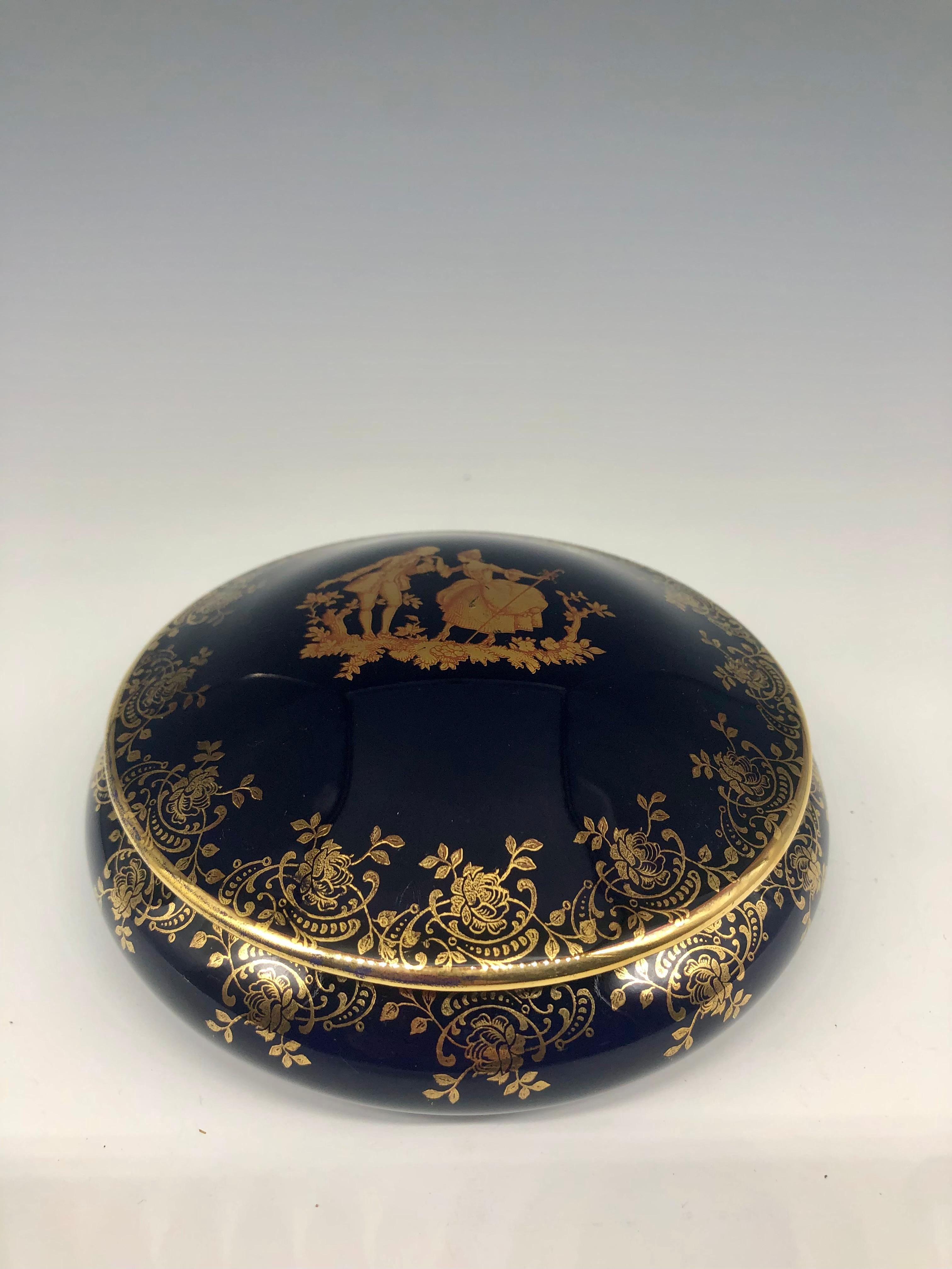 Elegante joyero vintage de porcelana azul cobalto con tapa y borde decorativo floral pintado a mano en dorado. La ilustración central representa a una pareja vestida al estilo de la época de Luis XVI, en la que el hombre besa la mano de una mujer