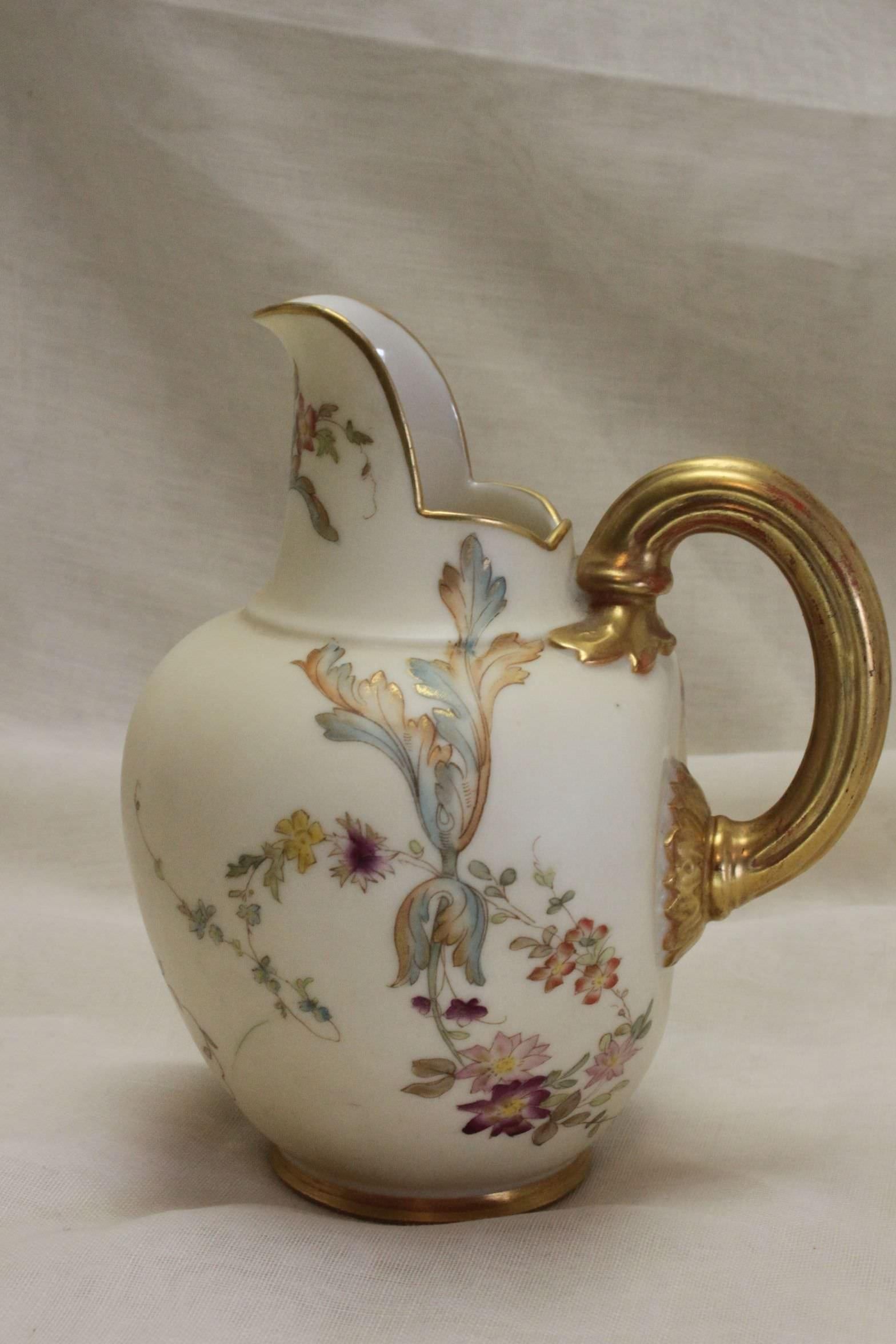 This small Royal Worcester jug is decorated with a rather unusual hand coloured and gilded pattern on a blush ivory ground. The pattern consists of scattered floral sprays that have been coloured using a delicate, restrained palette. The jug shape