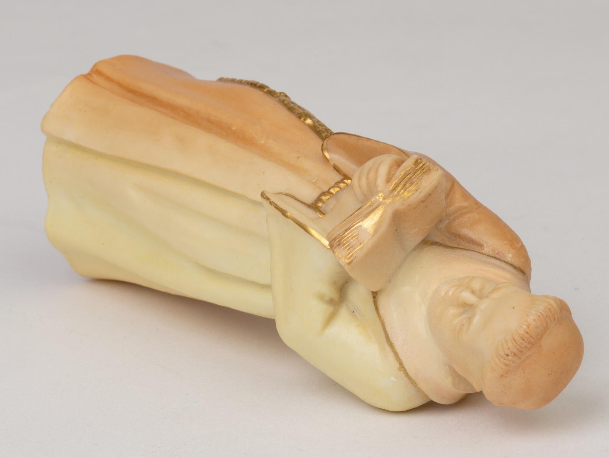 Un apagavelas antiguo de porcelana Royal Worcester, de muy fina factura, modelado como un monje leyendo una biblia y fechado en 1904. El apagavelas es representado como un monje vestido con túnica monástica atada a la cintura, que sostiene una