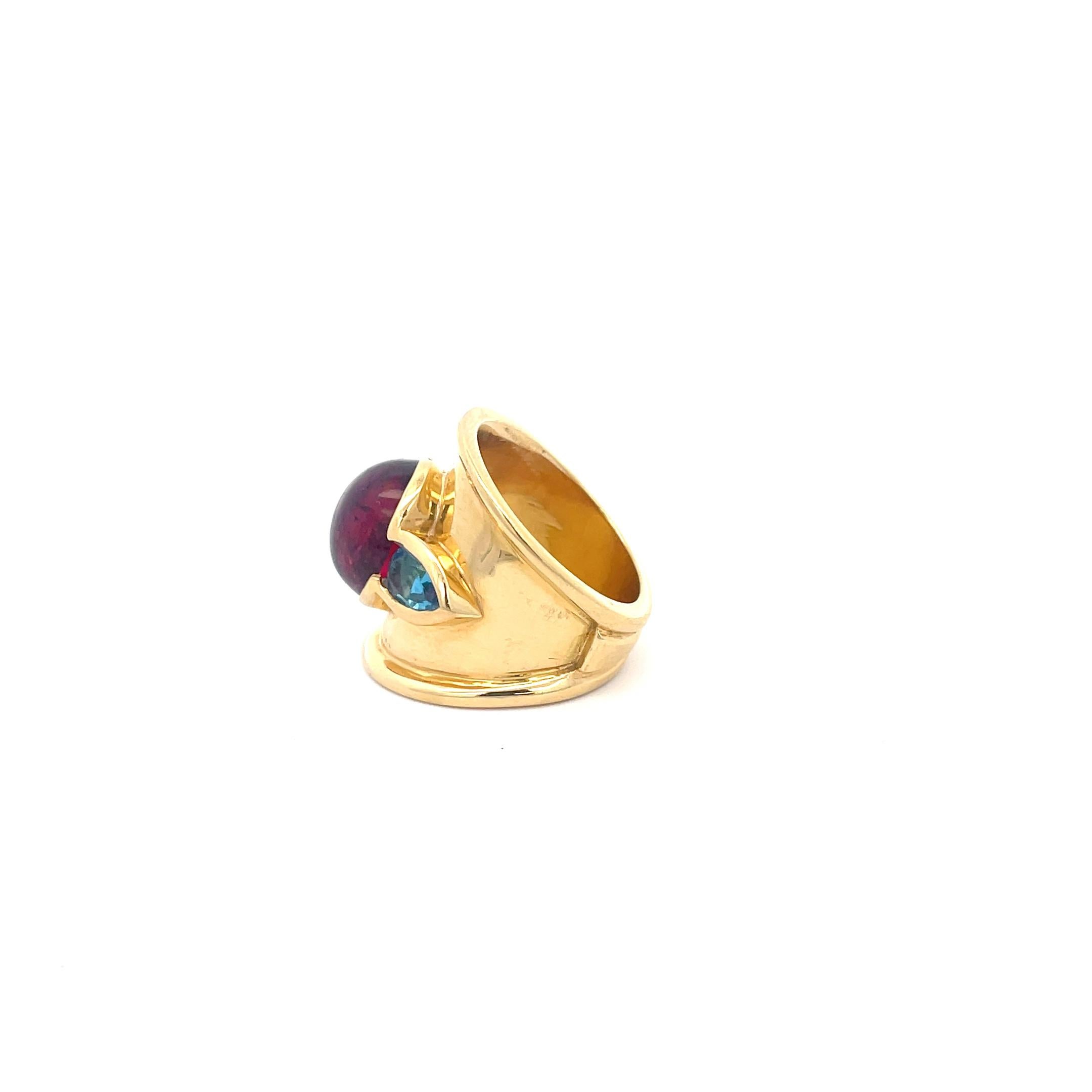 Rubellite and Blue Topaz Ring in 18K Yellow Gold. The ring features a cabochon rubellite (apprx 12.85mm x 10.50mm) and two pear shape blue topazes (apprx 6.90mm x 4.90mm). The ring is 22.75mm at widest point. Ring size 6.25. 
15.6 Grams