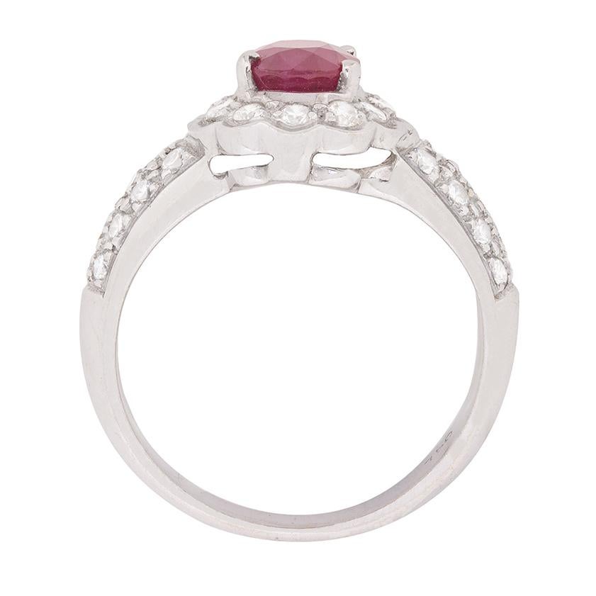 A halo of shimmering round brilliant cut diamonds envelops a bright oval-shaped ruby set in an open back four claw mounting at the centre of this sensational right hand ring. Pavé set shoulders lead to a wide shank, all in 18 carat white