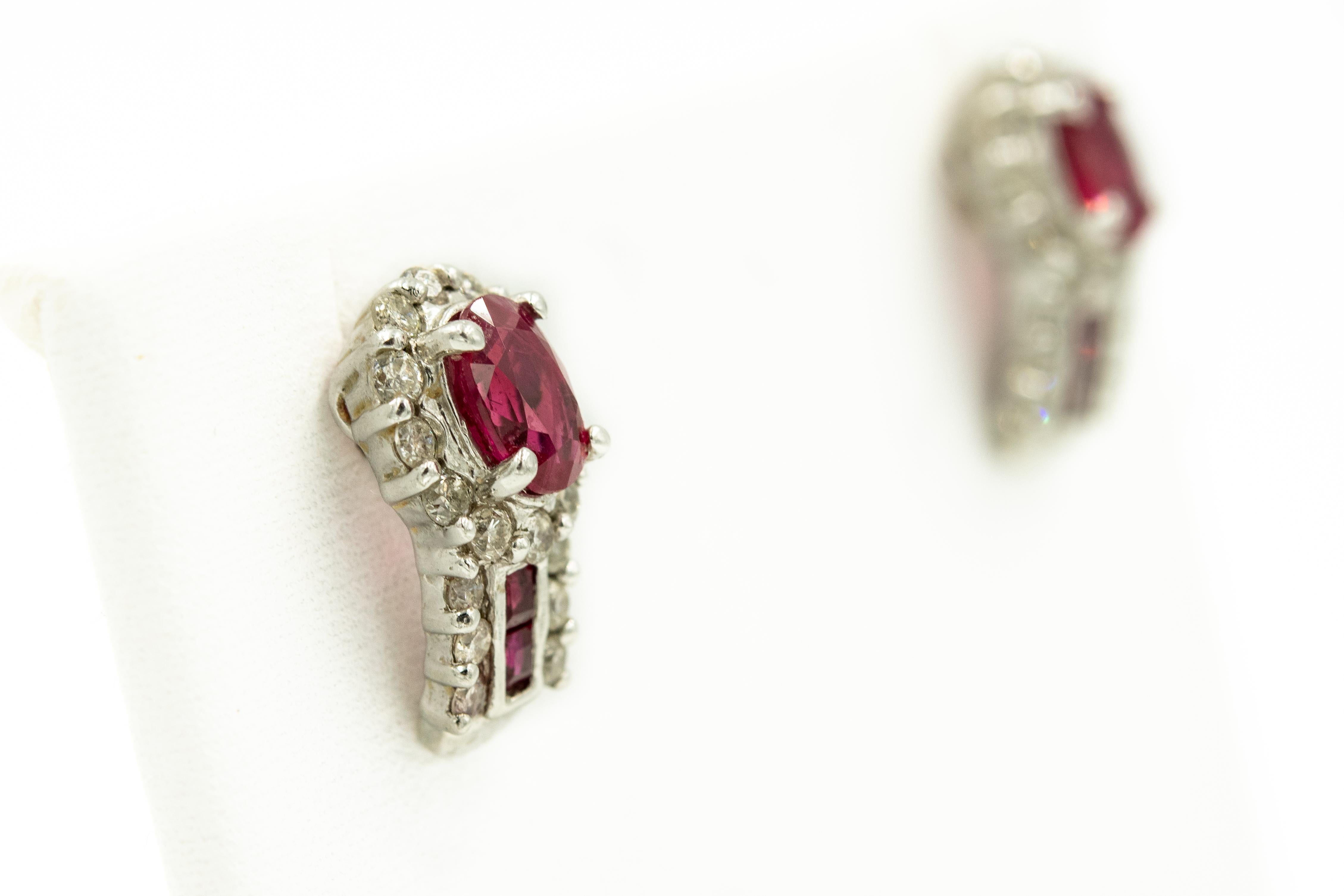 Small diamond and ruby white gold hoop type earrings featuring an oval ruby set in a diamond frame with a hopples area with channel set square rubies with three diamonds on either side.  The earrings are 18k white gold and the backs are 14k white