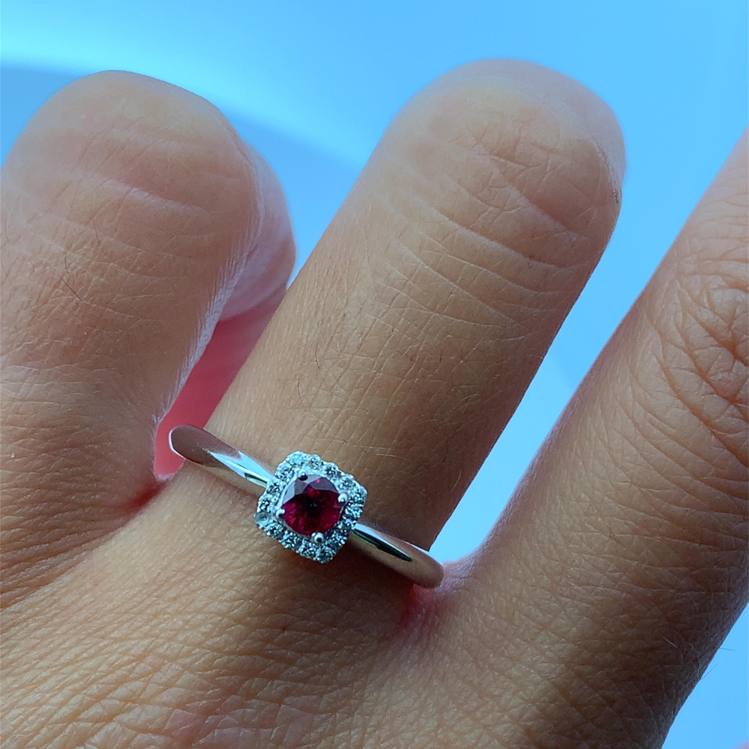 Squisitamente realizzato in lucente oro bianco 18 carati, questo anello solitario presenta un affascinante rubino a taglio rotondo e un'aureola di diamanti. Il taglio brillante dello 0,20ct con tonalità rosse  Il rubino Burma esalta il suo splendore