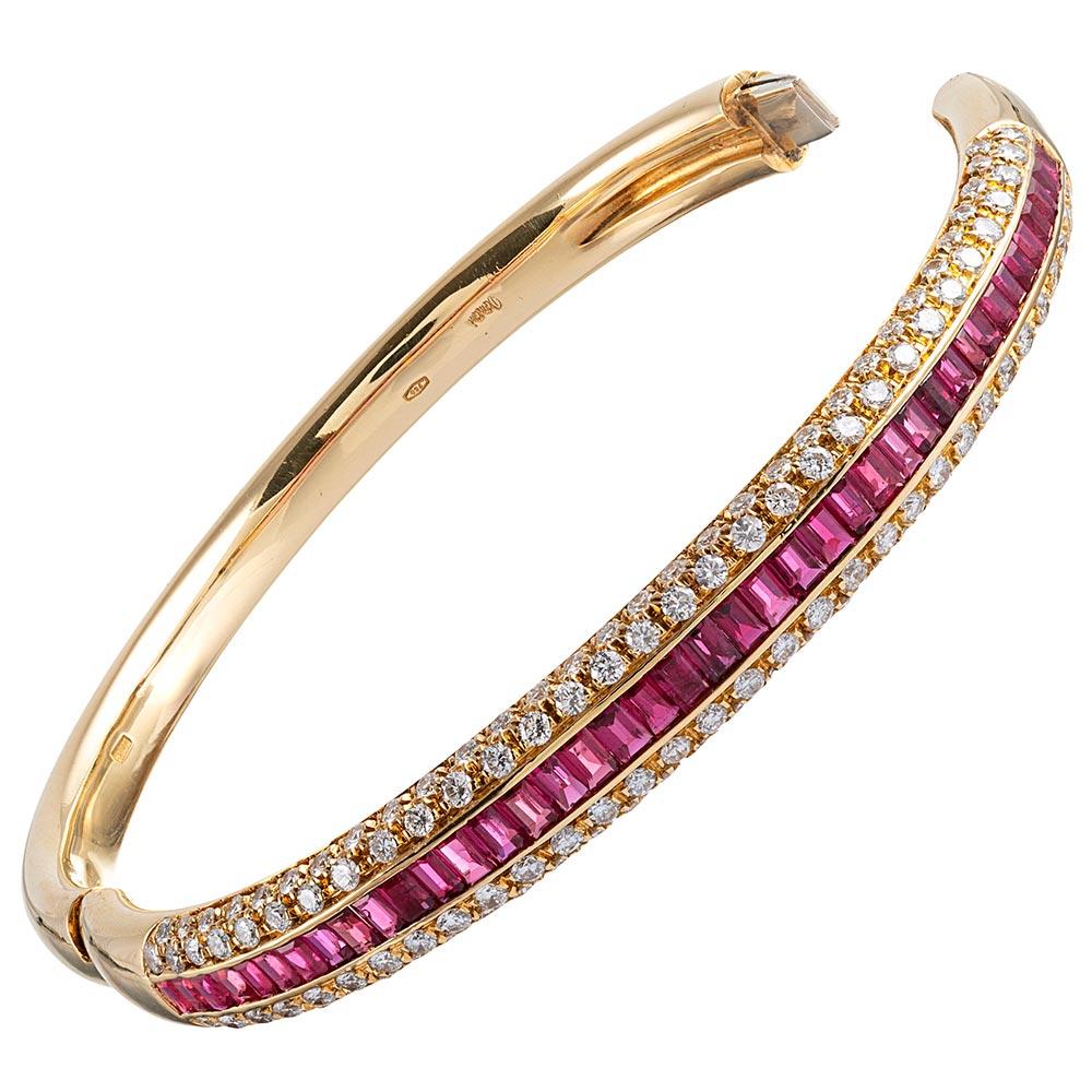 This substantial oval-shaped bangle made of 18 karat yellow gold is decorated with a single row of baguette rubies flanked by a staggered double row of brilliant white diamonds. The diamonds follow the natural curve of the top of the bracelet and