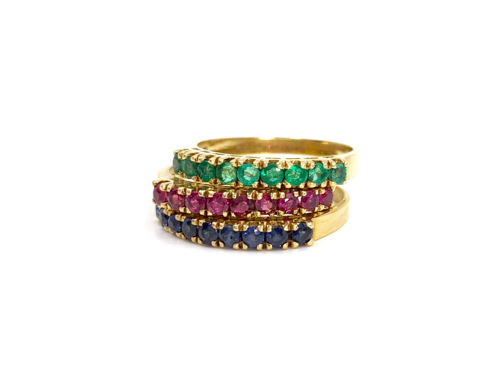 A set of three stackable 18 karat yellow gold 3mm band style rings, each featuring a straight row of 9 precious gemstones: rubies, blue sapphires and emeralds. Gemstones are securely set in four prong boxes. Approximate total weight of each ring is