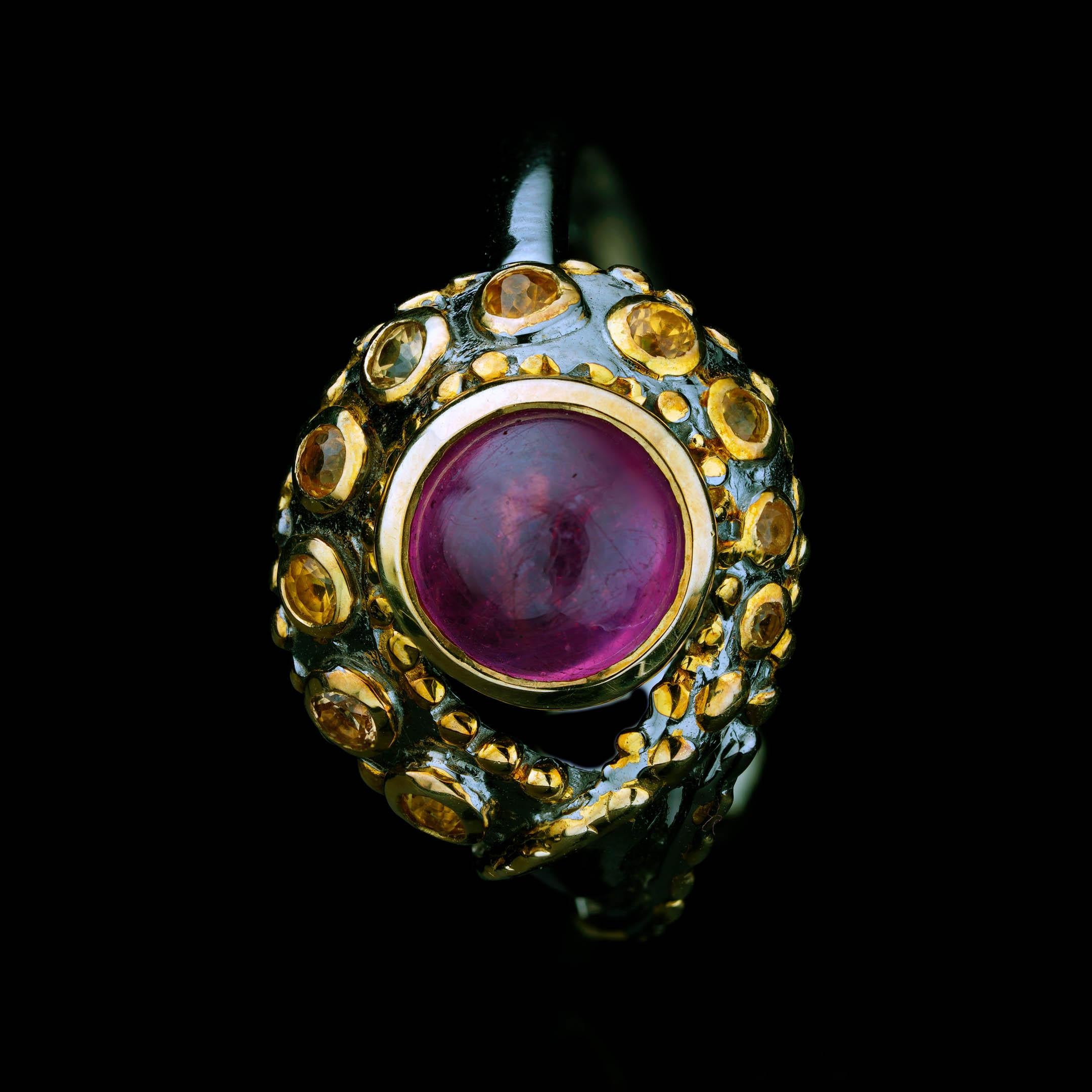 This artisan ring centers on a glowing natural cabochon Ruby, bezel-set in radiant 14K gold, and surrounded by a constellation of faceted yellow sapphires. The sculptural oxidized sterling silver setting flows like a curling tentacle, detailed with