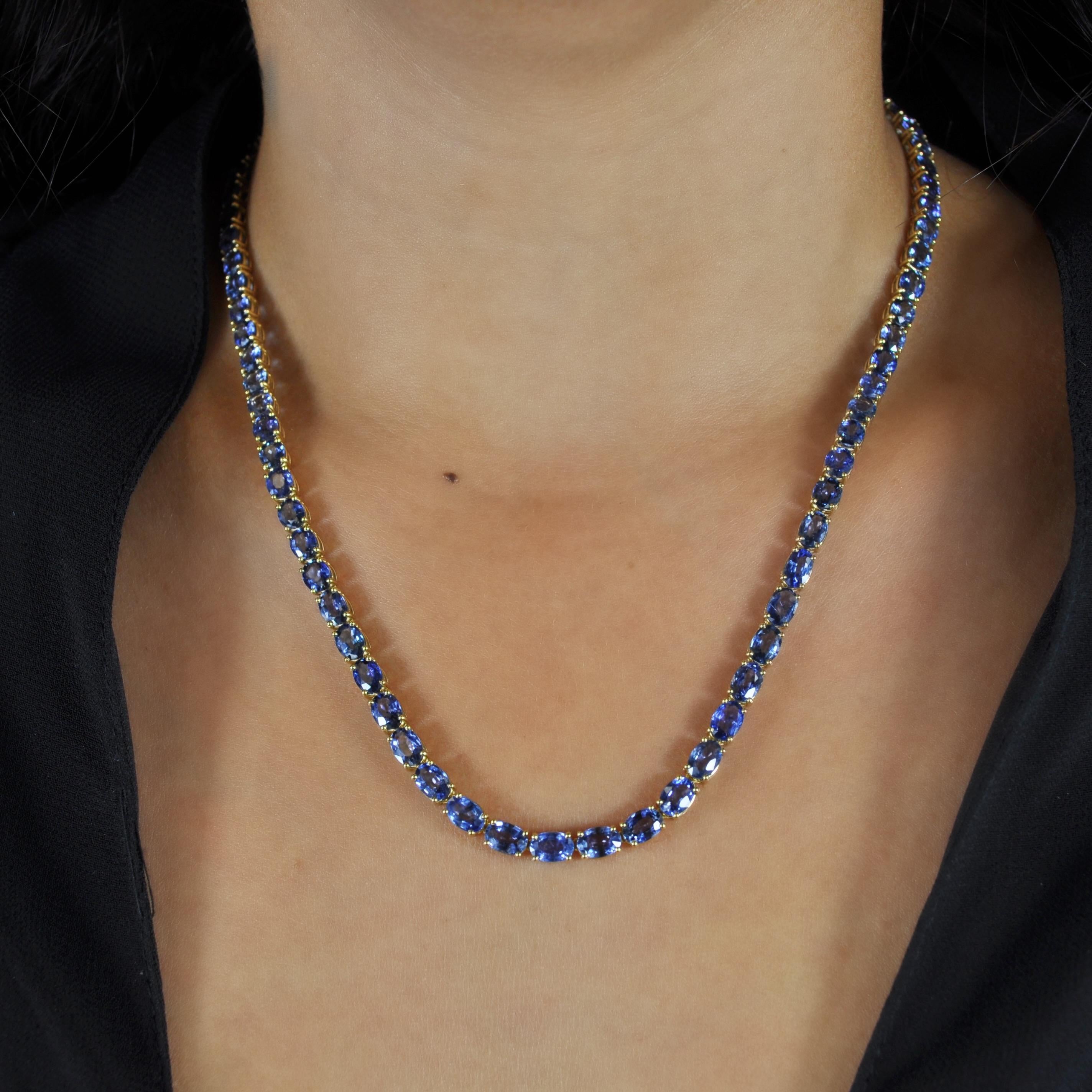 This 18k yellow gold link necklace features a total of 36.46 carats of oval-shaped blue sapphires encased in button prongs, with a box clasp and two figure-8 safety closures. It resembles the silhouette of a classic tennis bracelet but with a