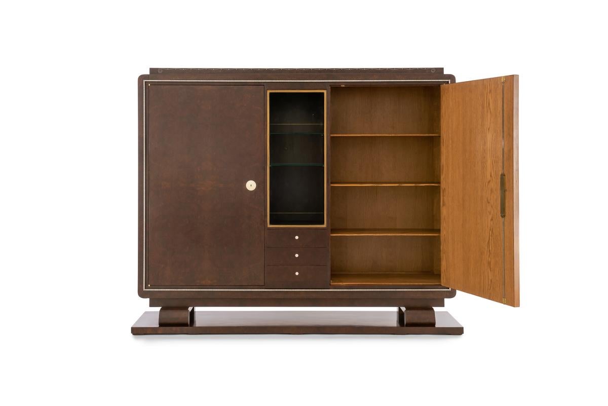 Important ‘Bibol’ Cabinet in Burl veneer on oak structure with ivory details. This display cabinet has two solid side doors, one glass door, and three drawers in the lower part. It has a lateral scroll foot resting on a rectangular base, with the