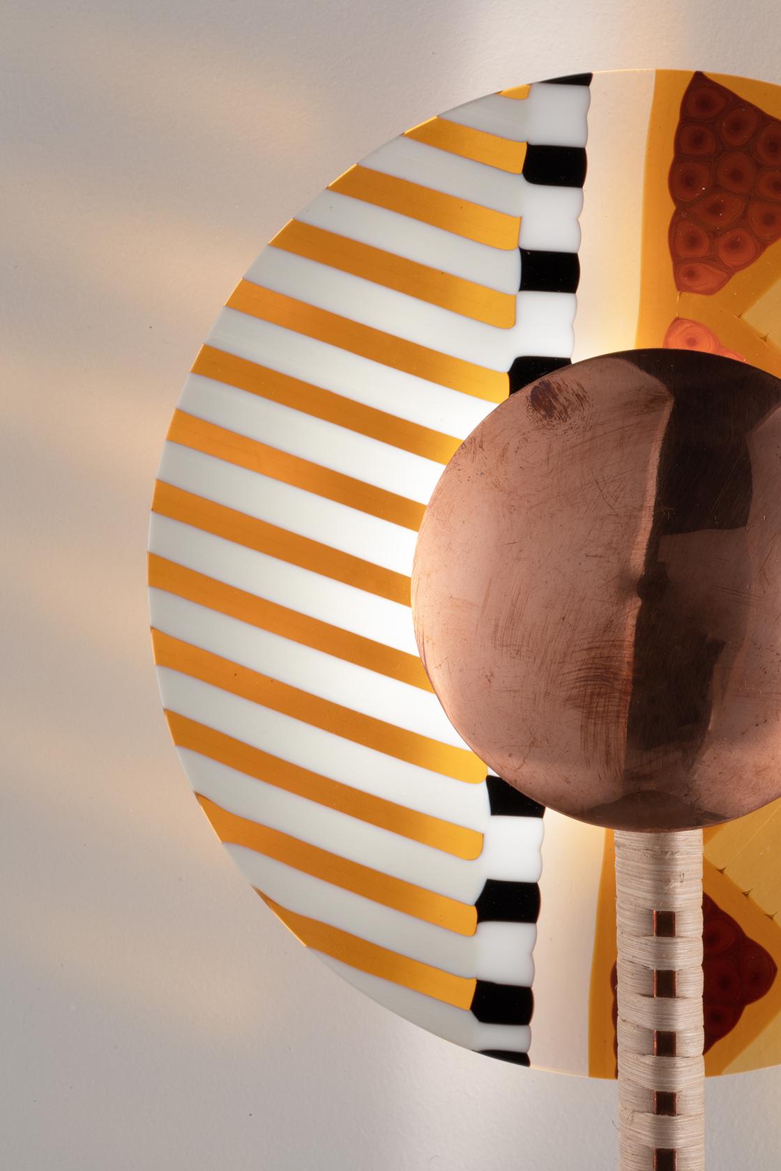 Lampada da parete a LED composta da un'asta in ottone Coates e da un disco di vetro colorato. La lampada è disponibile in quattro versioni, create da diverse combinazioni di elementi. L'asta è rivestita in rattan naturale intrecciato a mano con rame