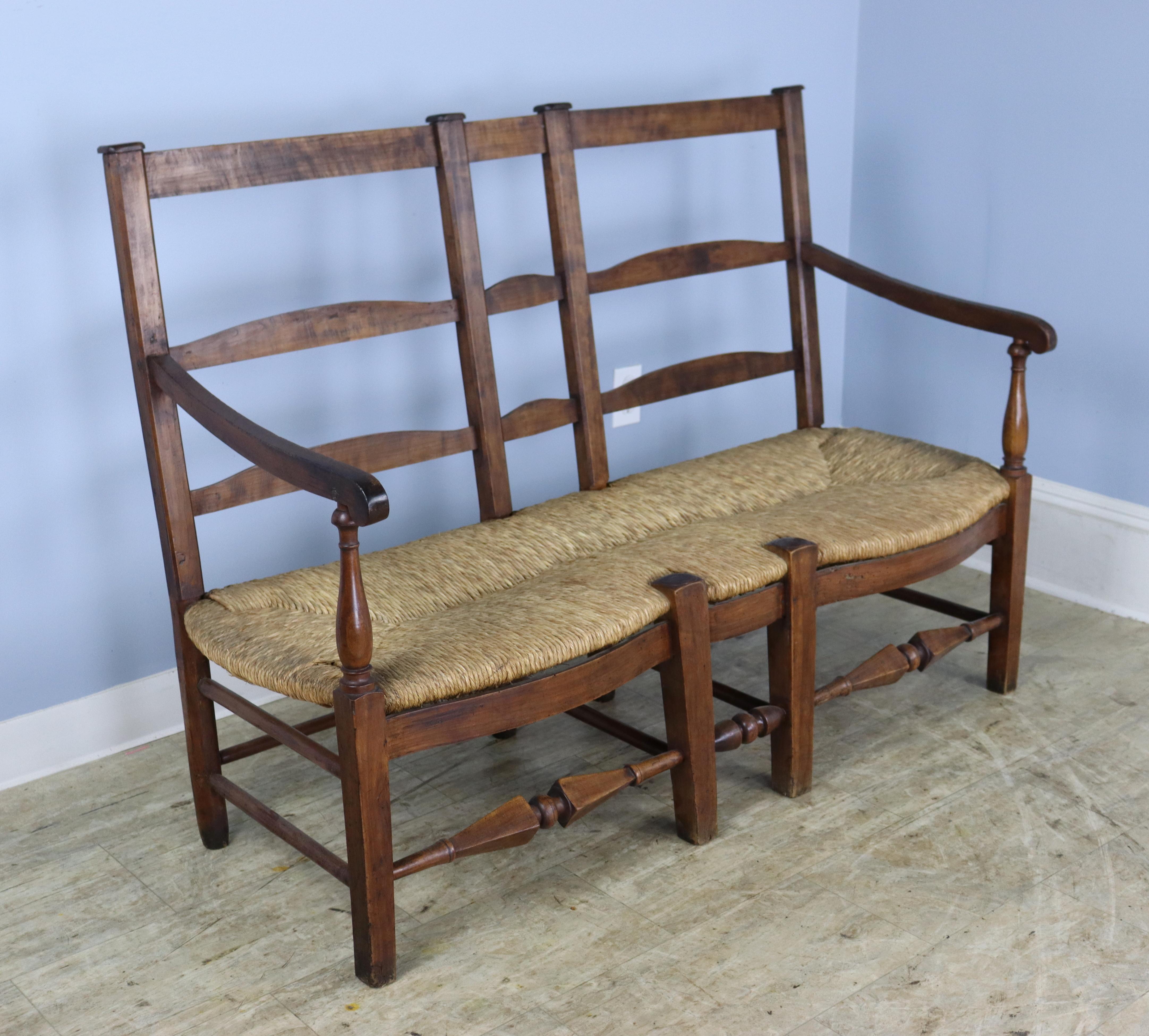 A rush seated French antique bench with Directoire era design elements, specifically the turning in the stretchers and the caps atop the back risers.  The cherry is a warm brown with good grain and some period appropriate wear, and the rush is in
