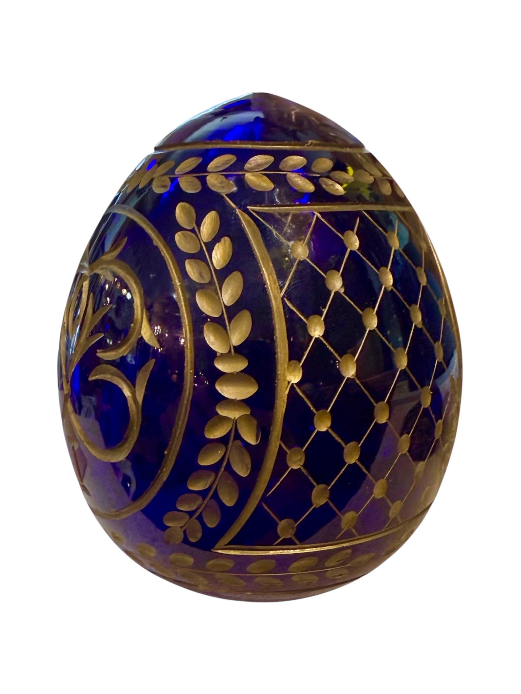 This exquisite Russian Fabergé-style egg is a striking homage to imperial opulence, crafted from rich cobalt blue glass and adorned with intricate gilded detailing. At the center of its design is a regal double-headed eagle, the enduring symbol of