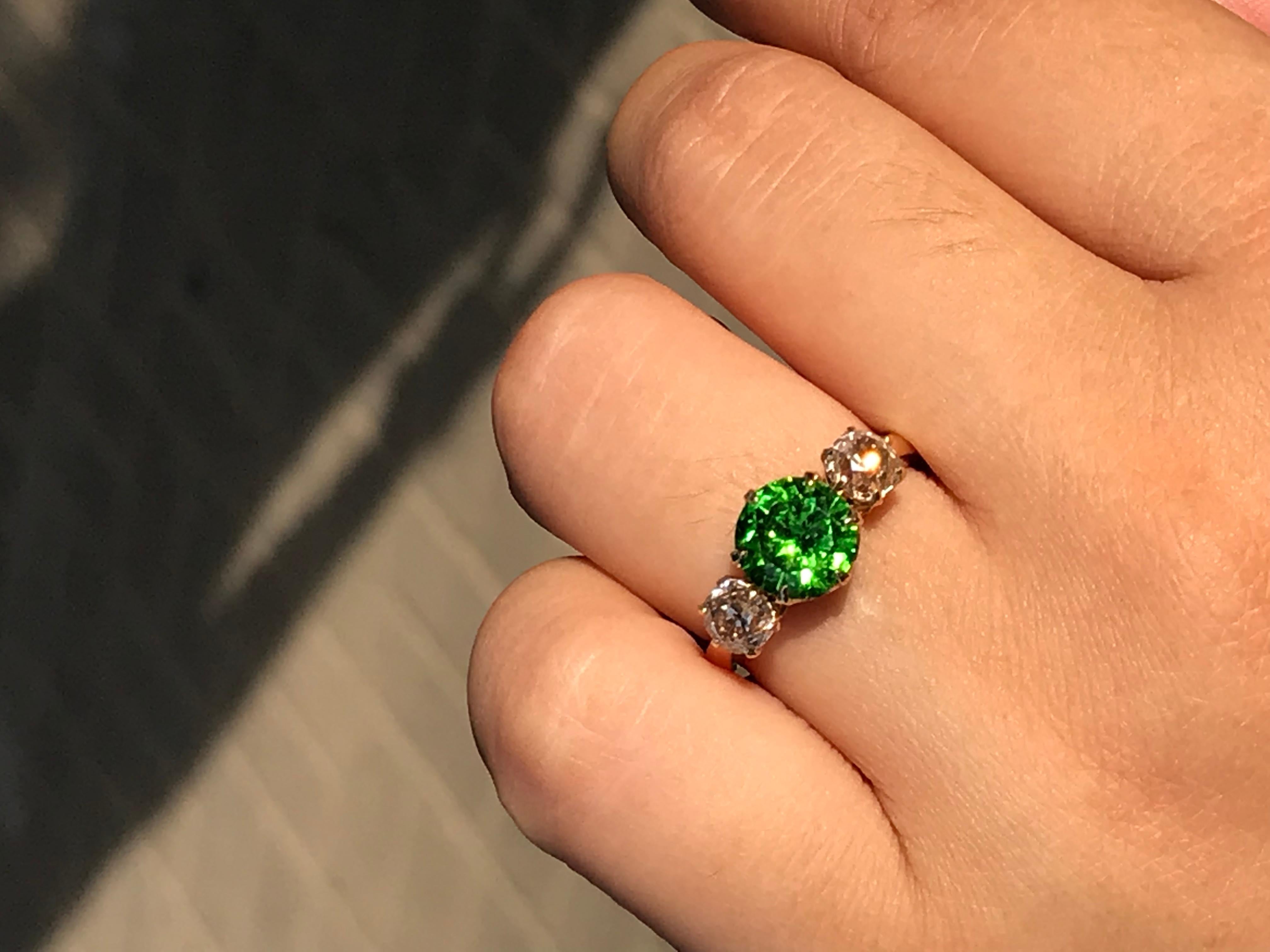 This stunning Russian Demantoid Garnet measuring 7.6mm in diameter is an incredibly vivid green colour. It is surrounded by two bright and lively diamonds which are M in colour and VS1/2-SI1 in clarity, each measuring at 4.6mm in diameter giving an